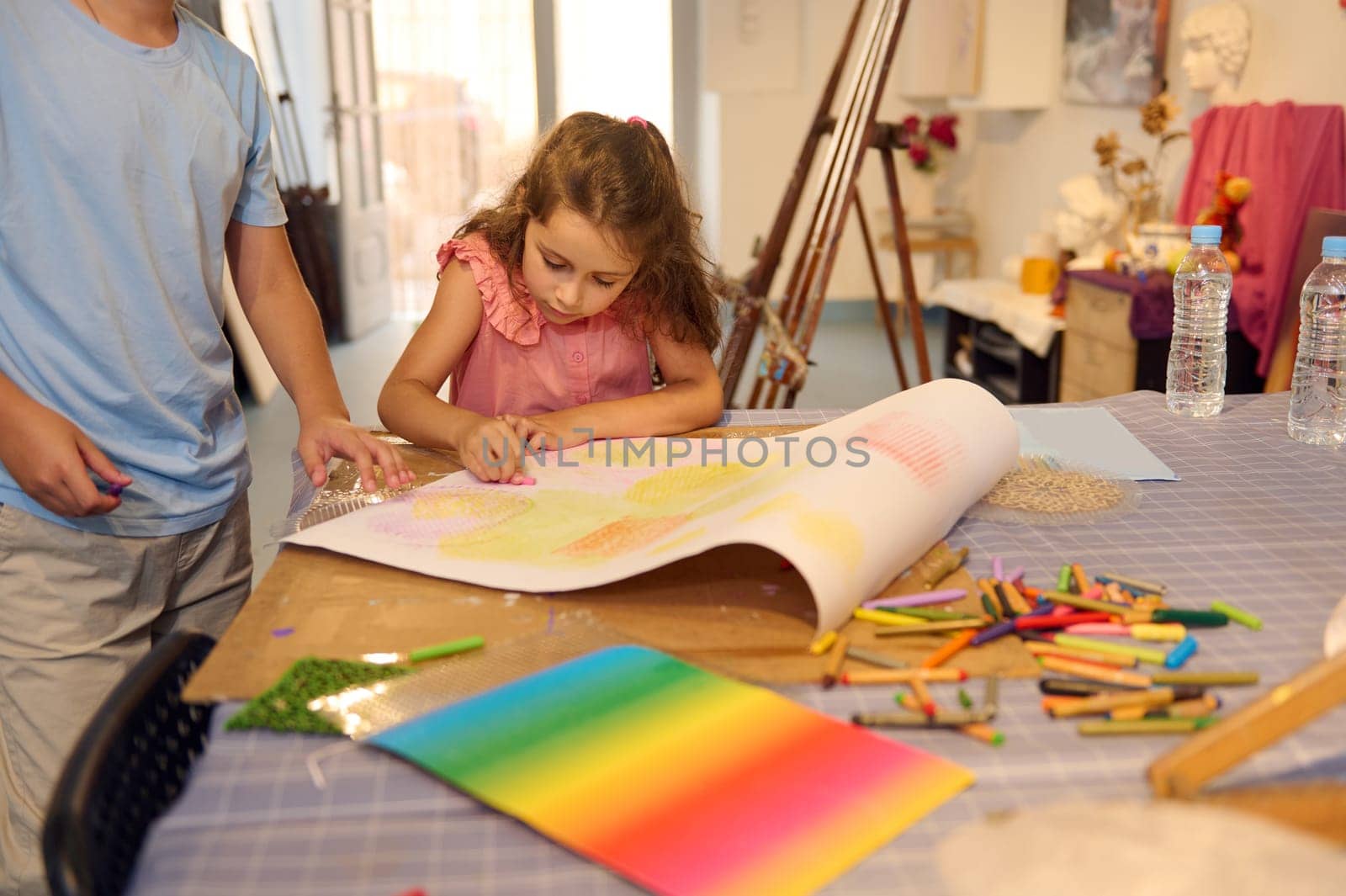 Talented inspired elementary age schoolgirl in pink dress, painting in the creative art school. Boy and girl learning drawing with pastels during visual art lesson. Artistic education. Fine art lesson