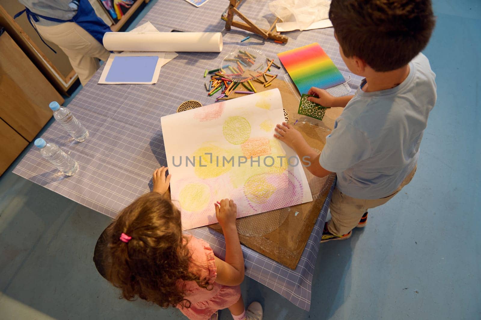 View from above of a teenage school boy and elementary age schoolgirl drawing on paper with pastel color pencils, creating a background for further during art class in the fine art school or gallery
