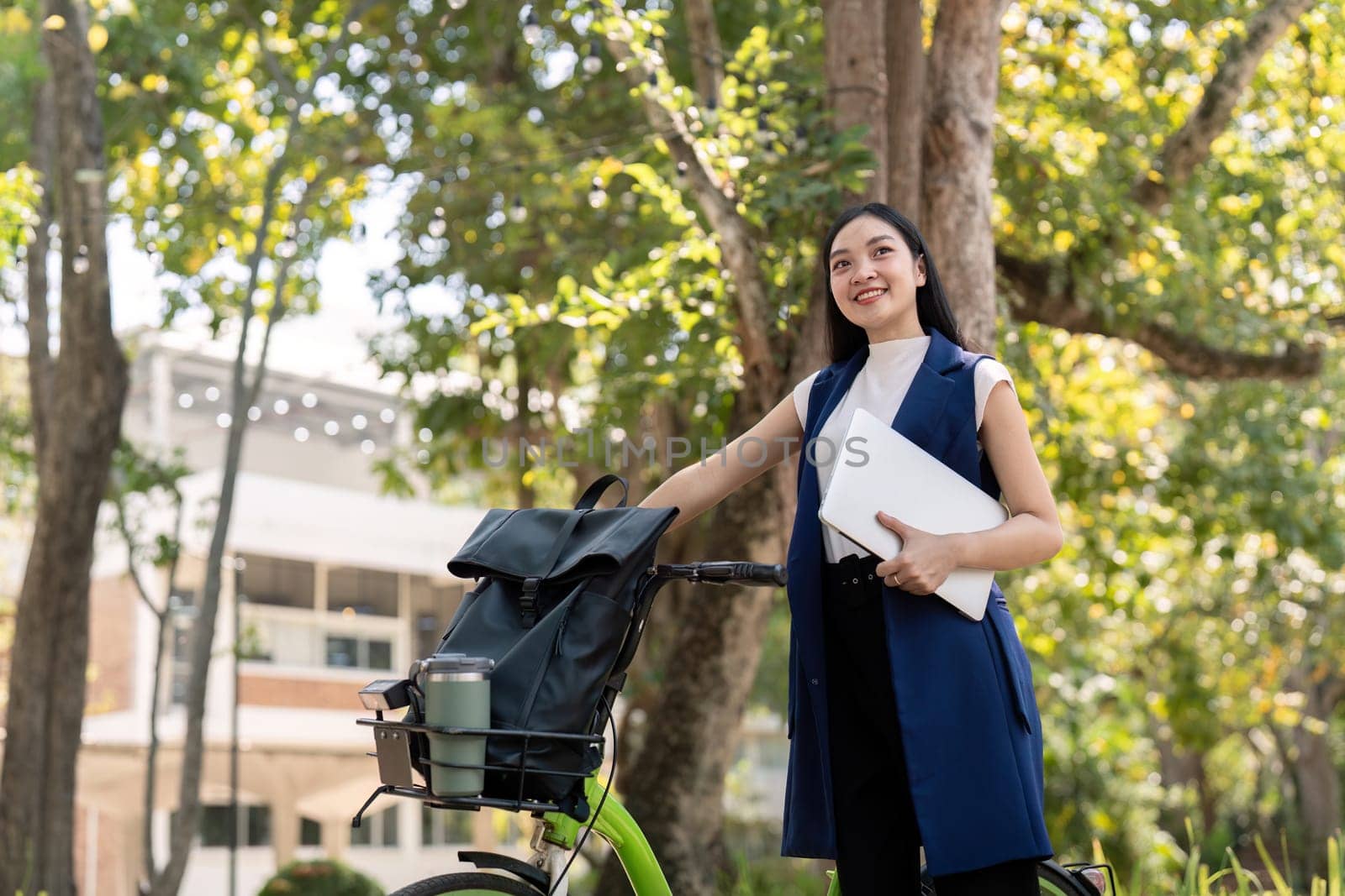 Asian businesswoman with bicycle holding a laptop and outside the office building. Woman commuting on bike go to work. Eco friendly vehicle, sustainable lifestyle concept.