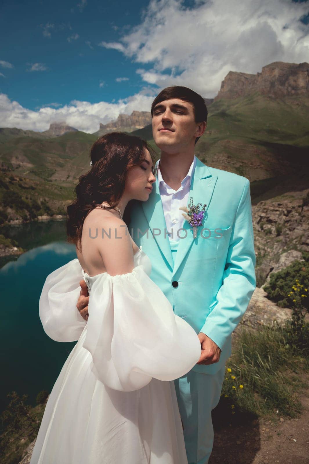 A beautiful wedding couple hugs tenderly against the backdrop of a mountain river and lake, the bride's long white dress.
