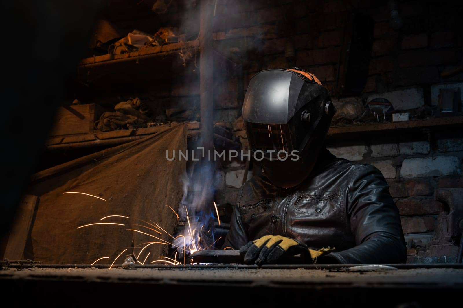 Young mechanic with a welding machine in an old dirty garage at night. Man in protection doing welding work, hobby by yanik88