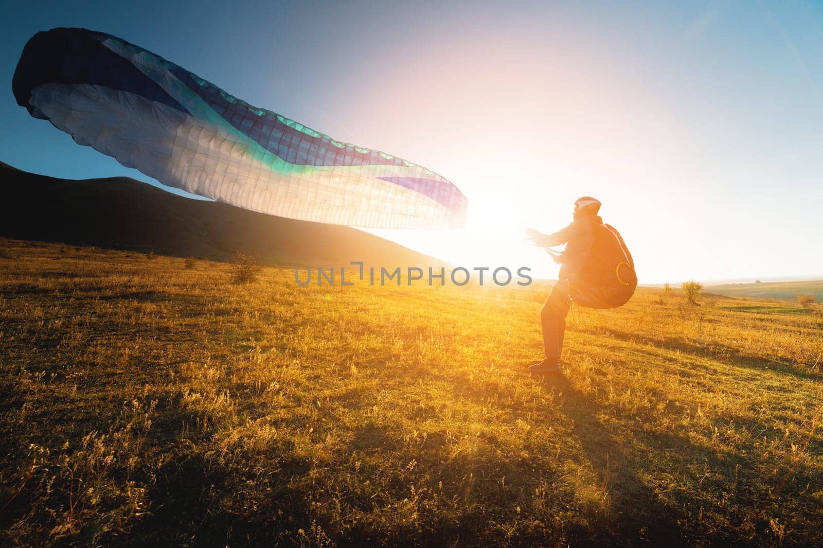 A paraglider with a blue parachute takes off. A male athlete stands on the field and lifts a paraglider into the air.