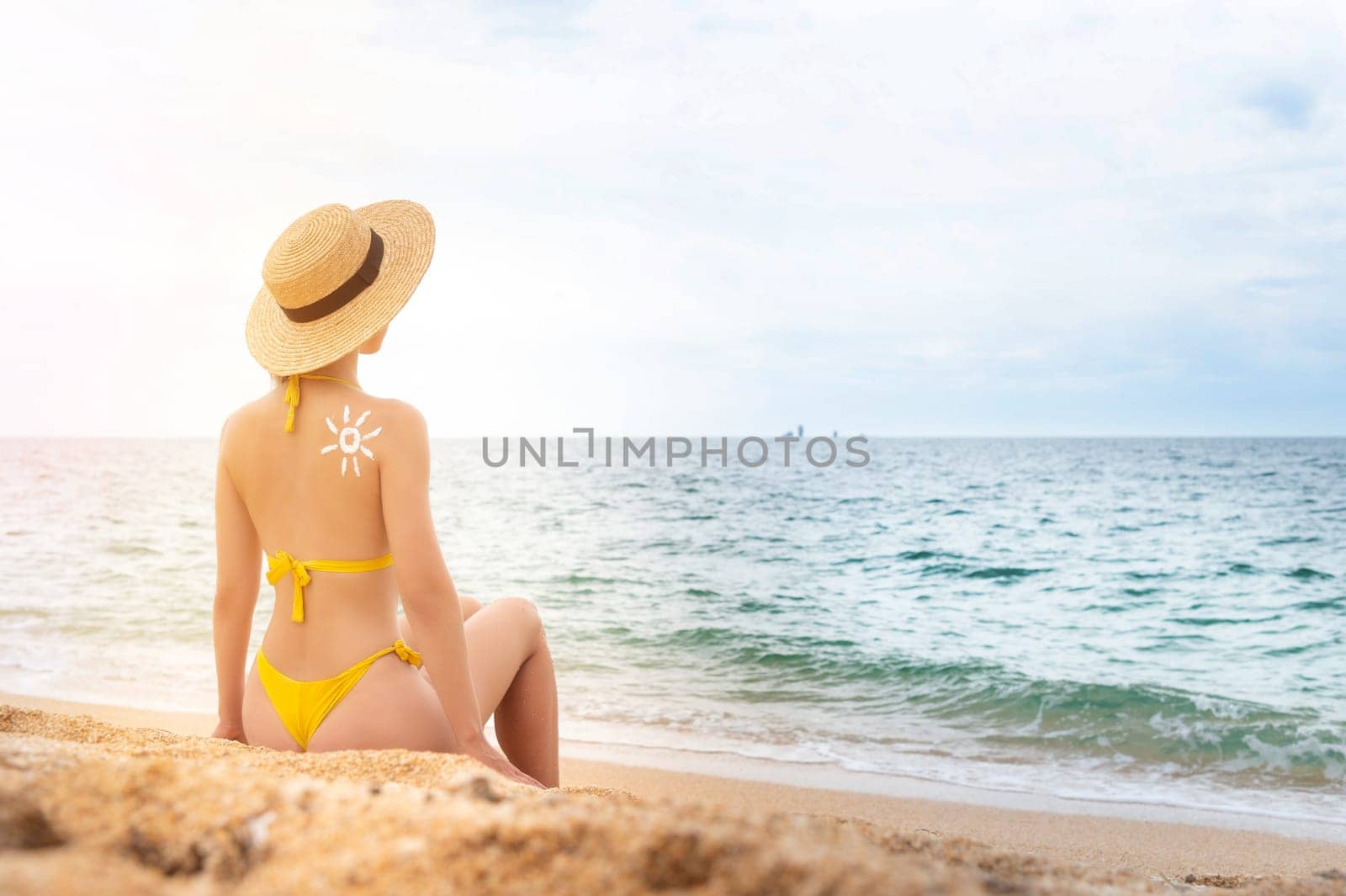 A young Caucasian woman in a swimsuit and a straw hat sits on the beach in front of the sea with her back in the frame. The sun on a woman's back is drawn with sunscreen.