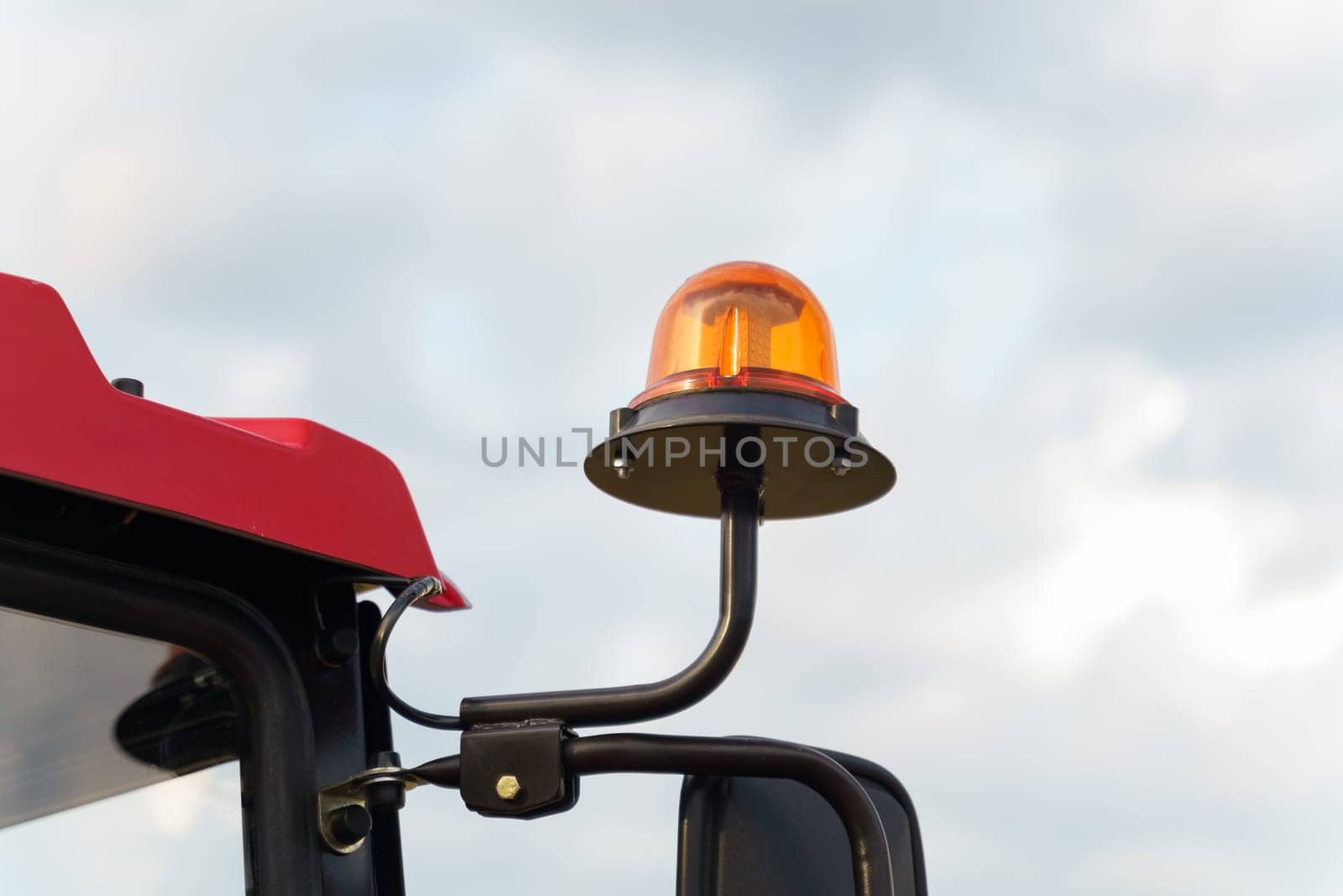 Orange warning beacon light mounted on the cab of a red farm tractor, symbolizing agricultural safety, captured against a backdrop of a partly cloudy sky.