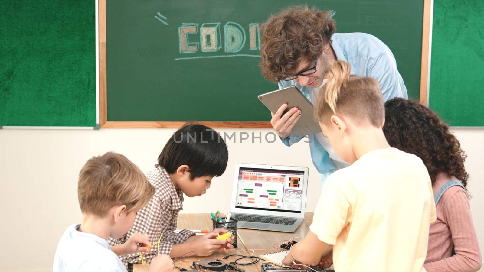 Group of diverse student working together on main board with laptop screen display engineering prompt written while teacher hold tablet and explain online software at STEM technology class. Pedagogy.