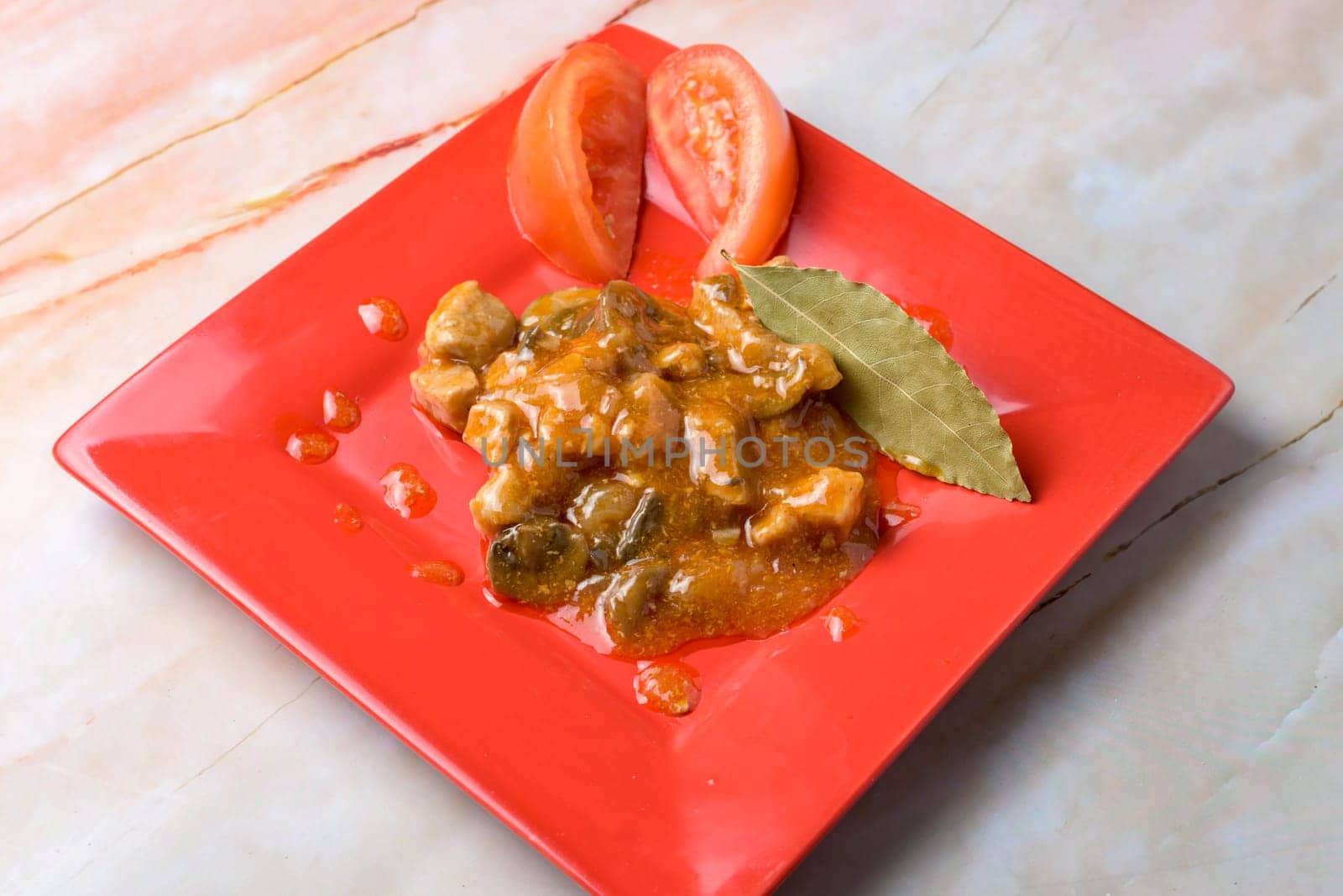 Pork stew with a bay leaf and tomato garnish on a red plate, marble background, typical food, typical mediterranean mallorcan cuisine typical from balearic islands mallorca, spain,