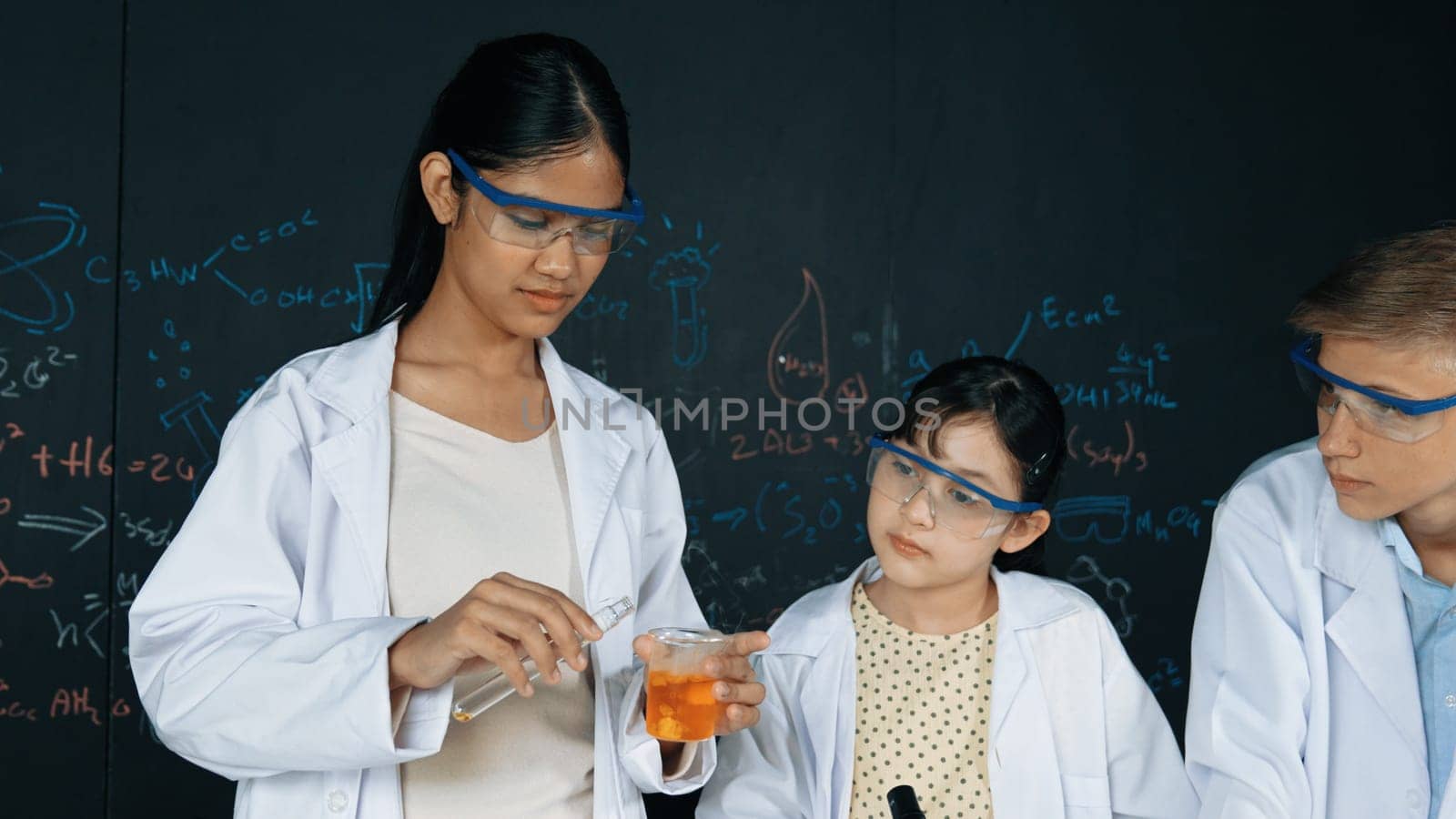 Smart girl pour colored solution in to beaker while diverse student excited about doing experiment. Professional scientist prepare for doing experiment at blackboard with chemical theory. Edification.