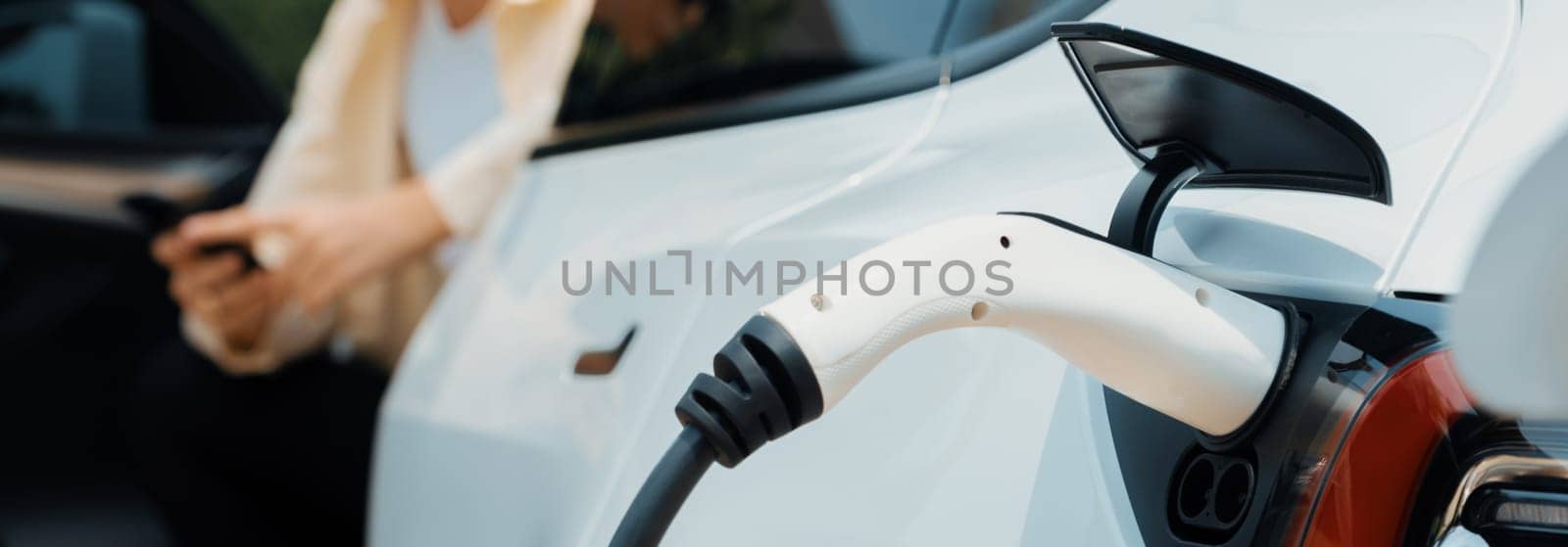 Young woman use smartphone to pay for electricity at public EV car charging station green city park. Modern environmental and sustainable urban lifestyle with EV vehicle. Panorama Expedient