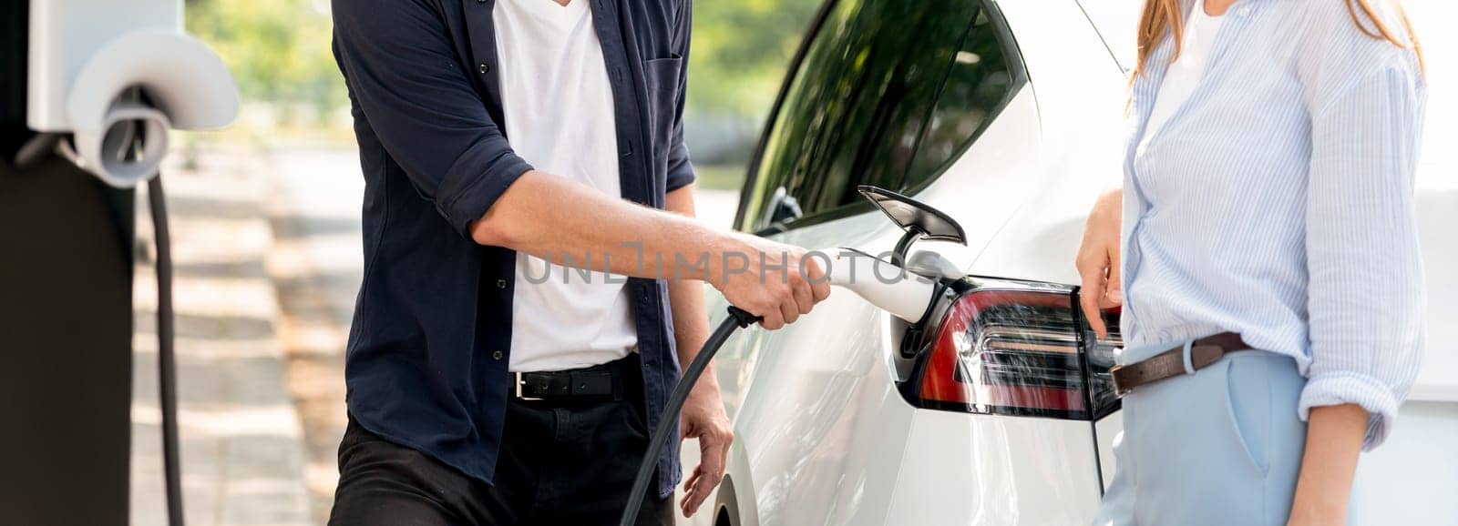 Focused EV car recharging electricity for battery on blurred background of lovey couple ruing their road trip travel by eco friendly electric car in national park and greenery forest on holiday. Exalt