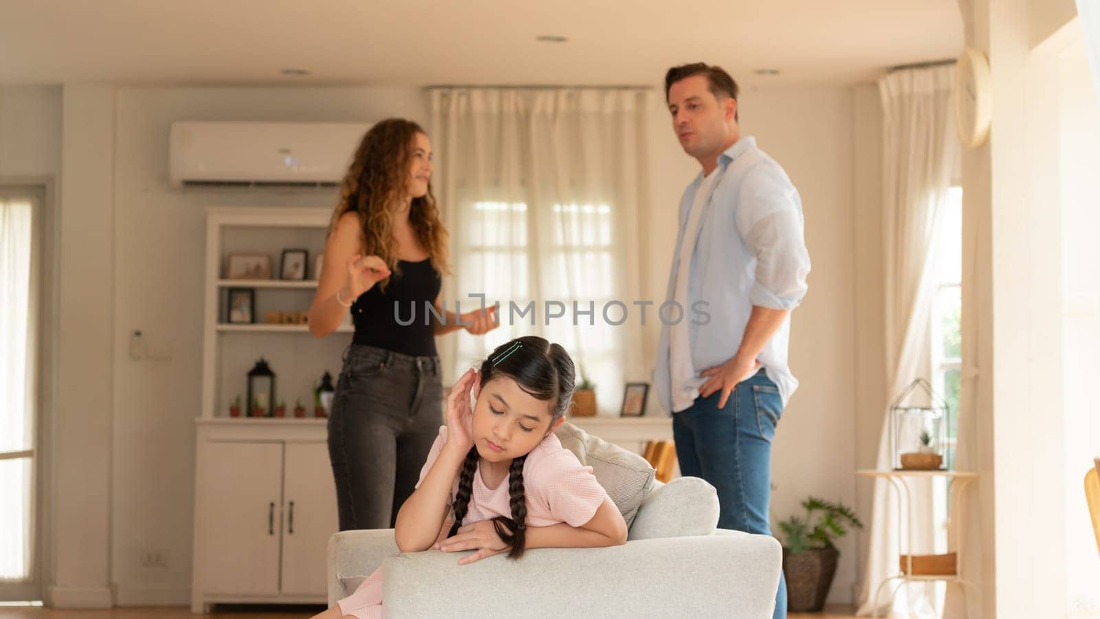 Annoyed and unhappy young girl sitting on sofa trapped in middle of tension by her parent argument in living room. Unhealthy domestic lifestyle and traumatic childhood develop to depression.Synchronos