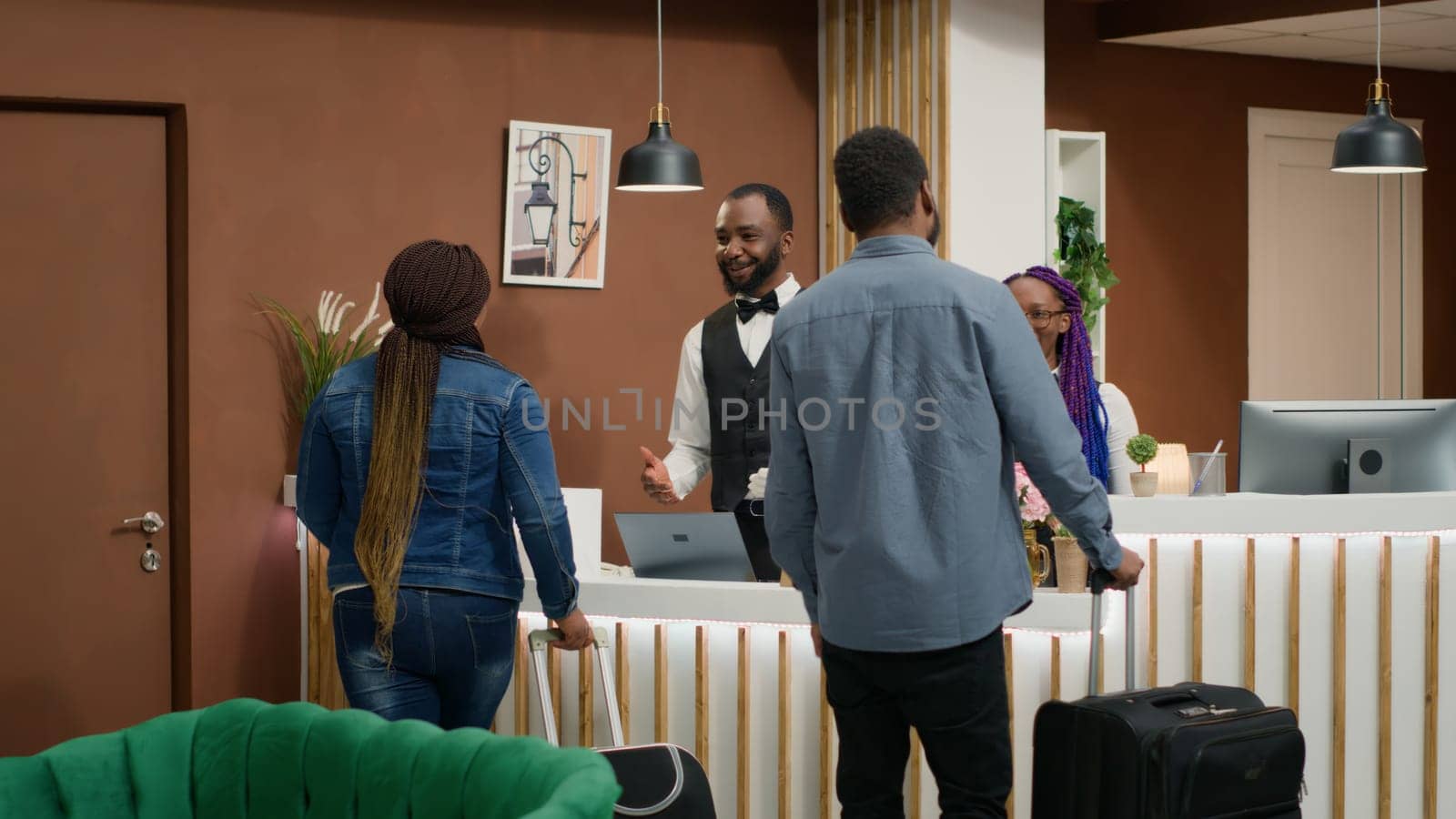 Wife and husband arrive at resort, discussing with receptionist to about room booking and registration. People entering lounge area reception with luggage, honeymoon trip. Tripod shot.