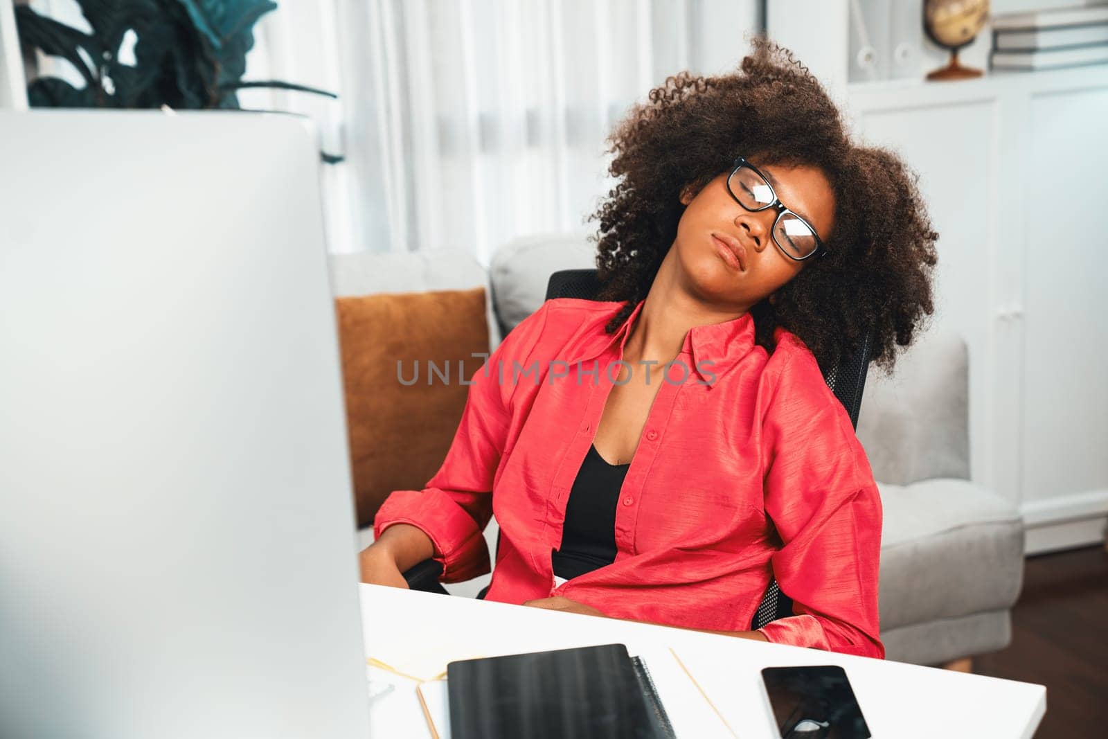 Young African woman taking a nap with dress up colored suit, leaning on office chair at workplace, waring glasses while sleeping. Concept of being exhausted of working project job. Tastemaker.