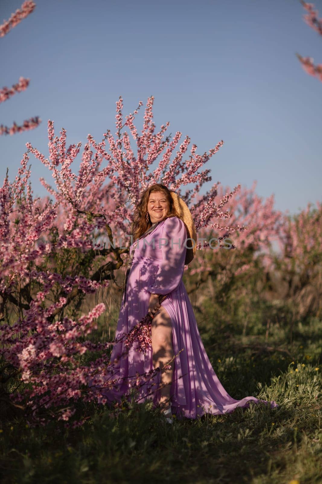 Woman blooming peach orchard. Against the backdrop of a picturesque peach orchard, a woman in a long pink dress and hat enjoys a peaceful walk in the park, surrounded by the beauty of nature