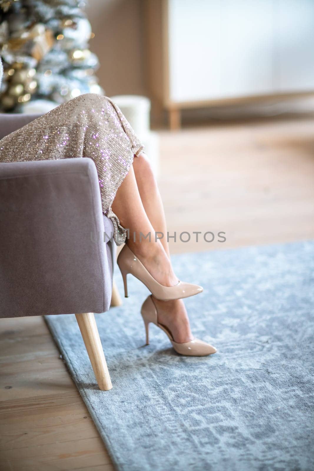 Women's legs. Cropped close-up photo of healthy beautiful elegant female legs in beige high-heeled shoes, woman sitting in a chair on the background of a Christmas tree with gifts by Matiunina