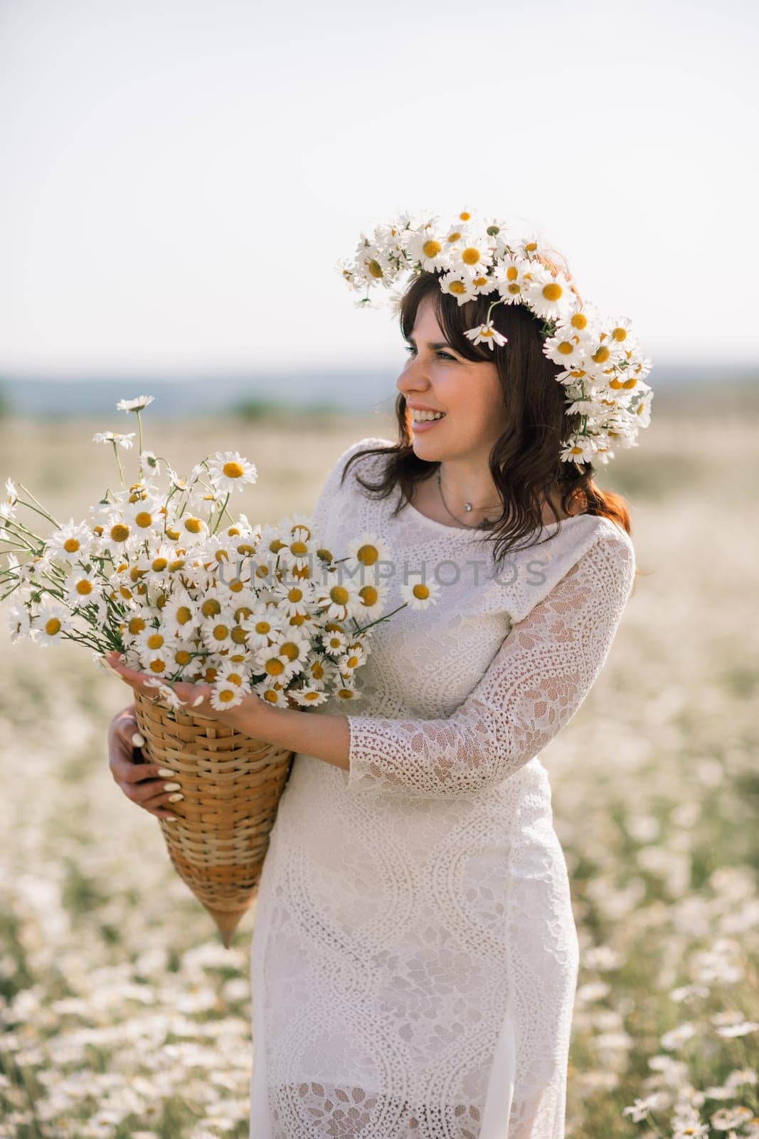 Happy woman in a field of daisies with a wreath of wildflowers on her head. woman in a white dress in a field of white flowers. Charming woman with a bouquet of daisies, tender summer photo.