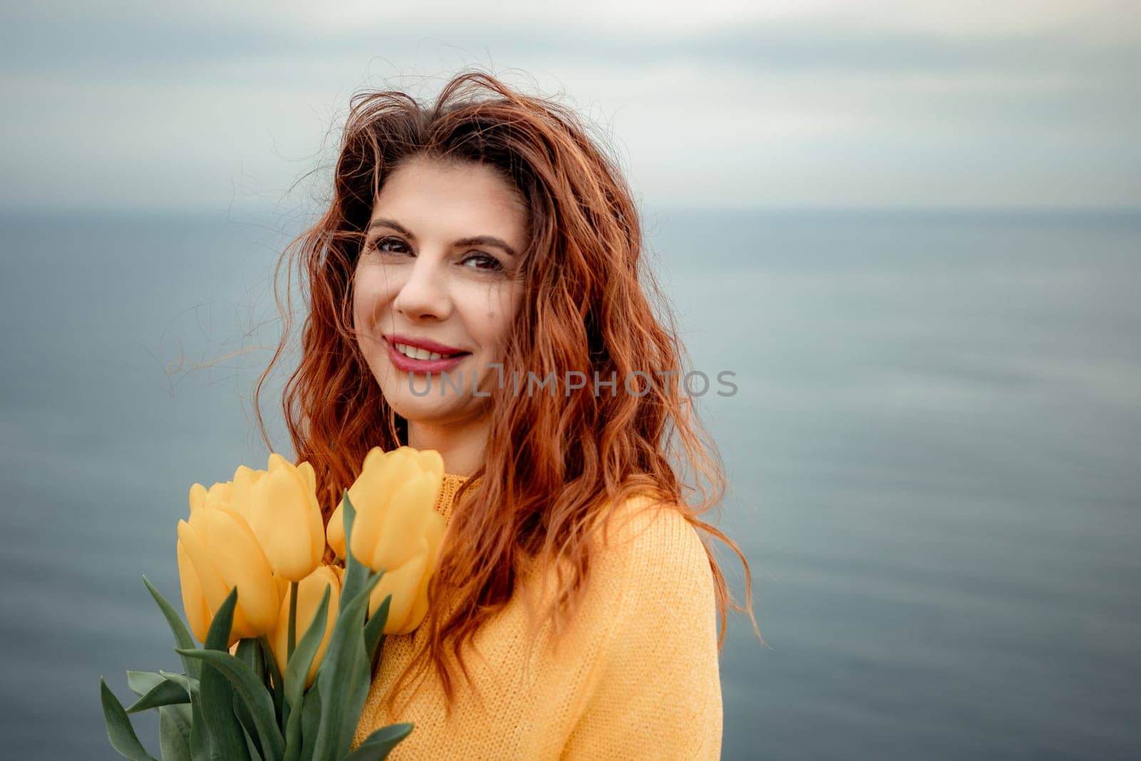 Portrait of a happy woman with hair flying in the wind against the backdrop of mountains and sea. Holding a bouquet of yellow tulips in her hands, wearing a yellow sweater.