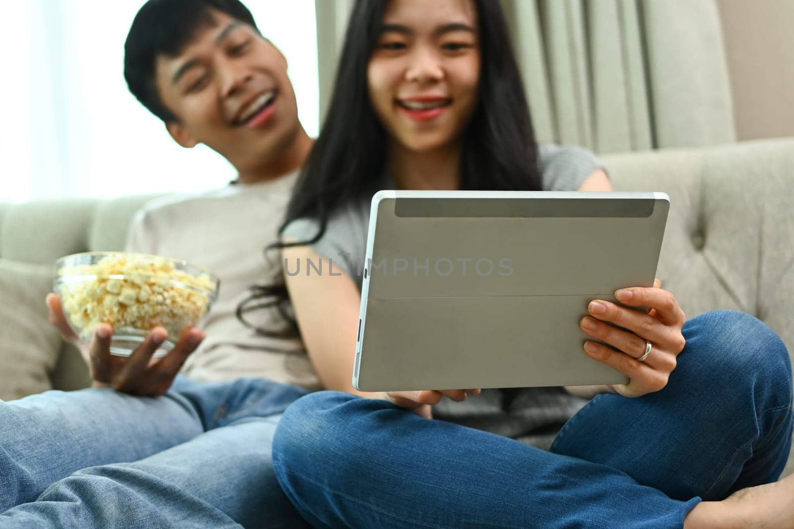Smiling young couple eating pop corn and watching movie on digital tablet in living room.