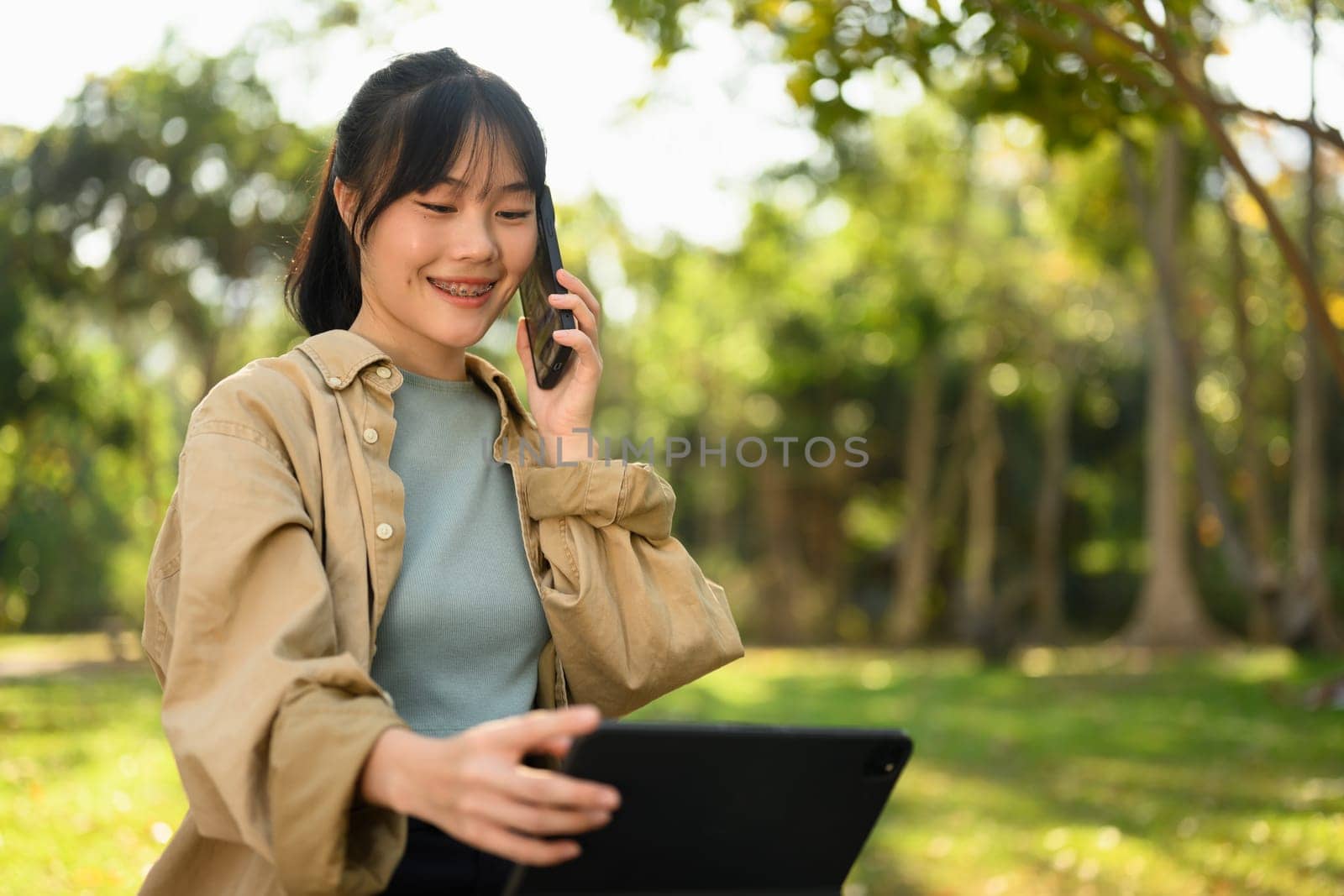 Smiling young woman talking on mobile phone and using digital tablet in the park on sunny day.