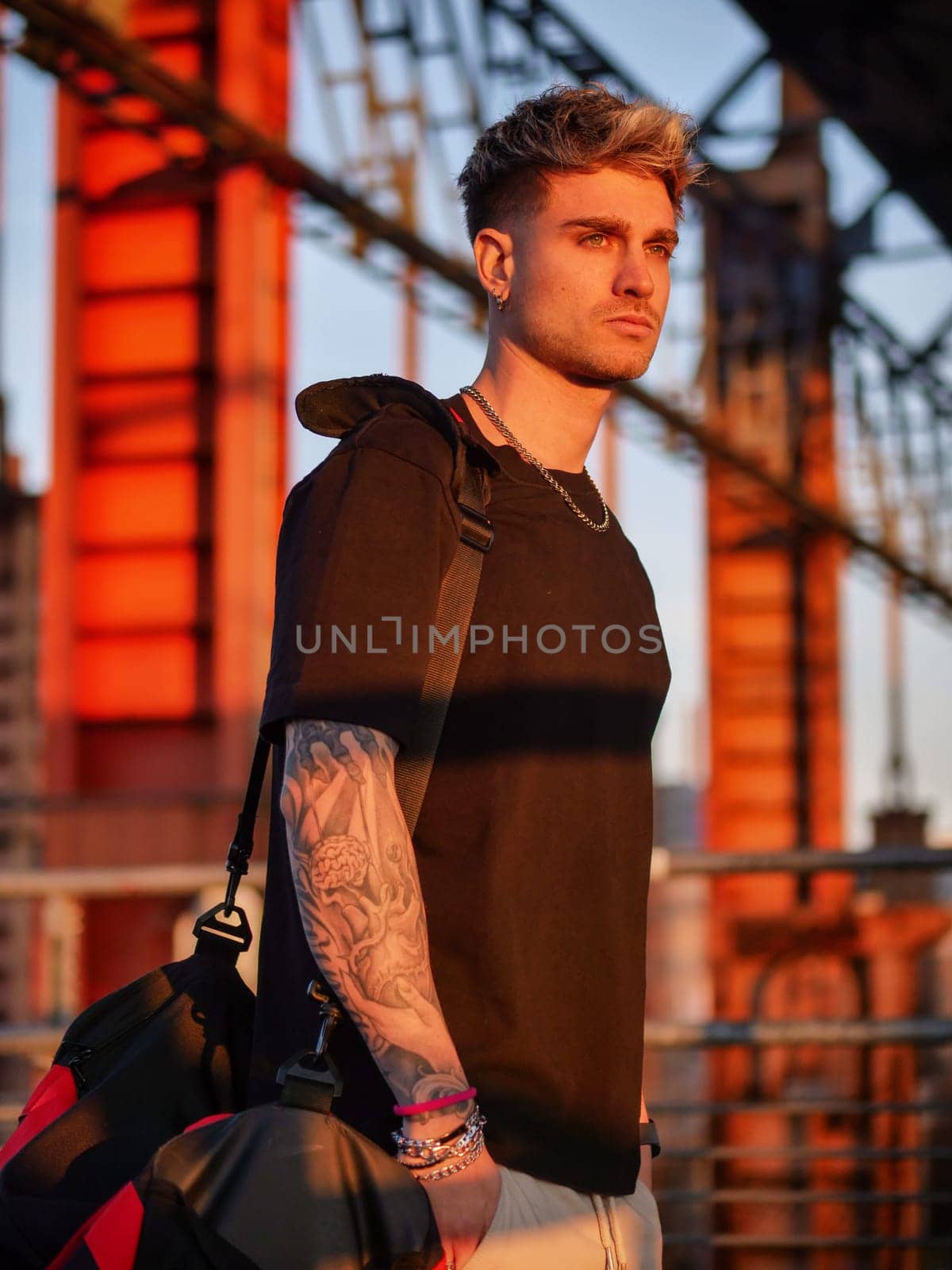 A man with a visible tattoo on his arm stands confidently in front of a modern building. He gazes ahead, creating a striking contrast between his edgy appearance and the sleek architectural backdrop.