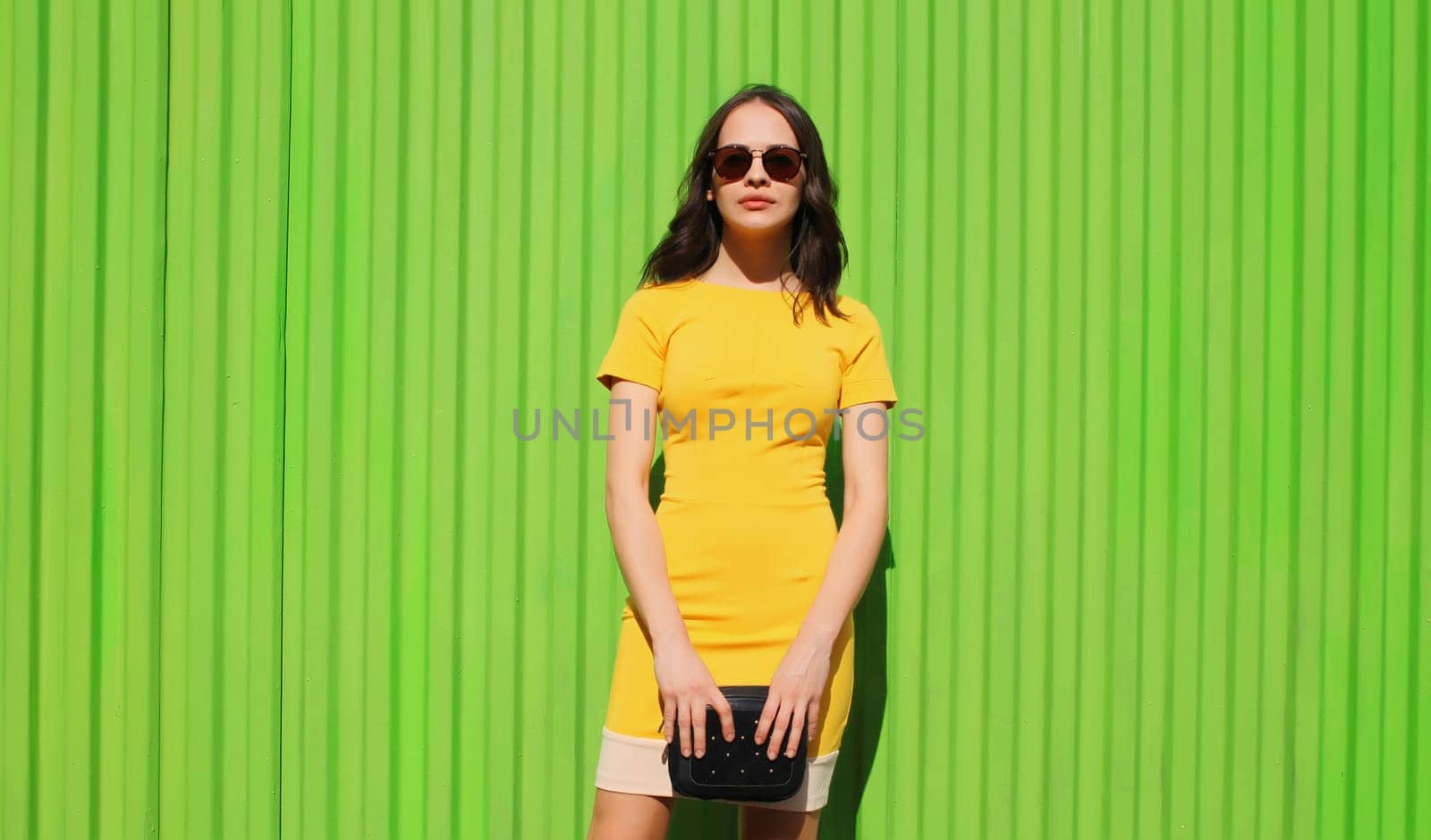 Beautiful young brunette woman holding handbag clutch posing in yellow dress on city street