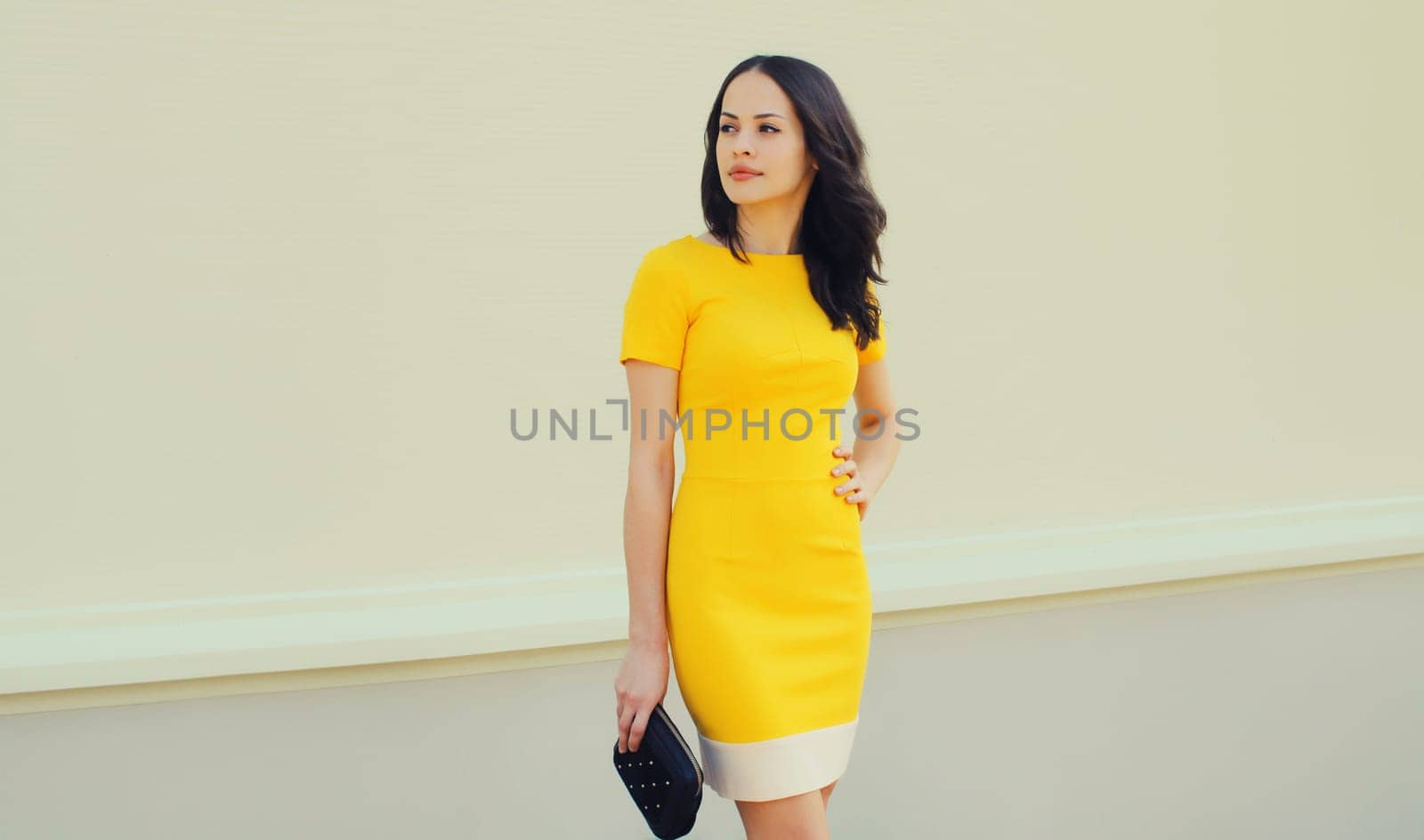 Beautiful young brunette woman holding handbag clutch posing in yellow dress on city street