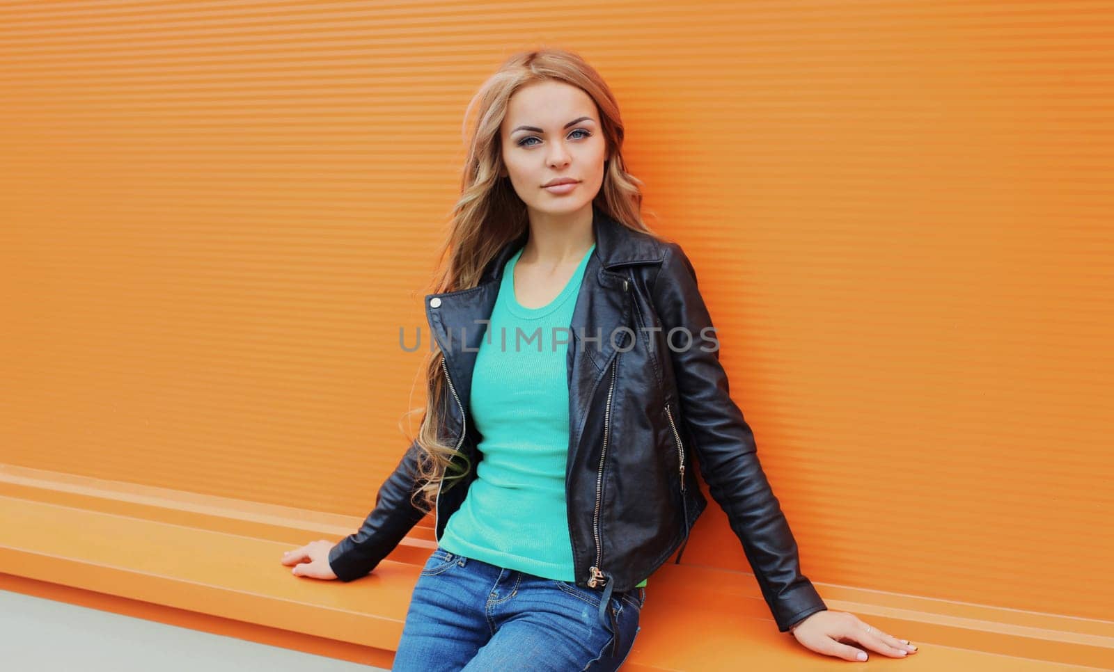 Portrait of beautiful young blonde woman posing in black leather jacket looking at camera on colorful orange background