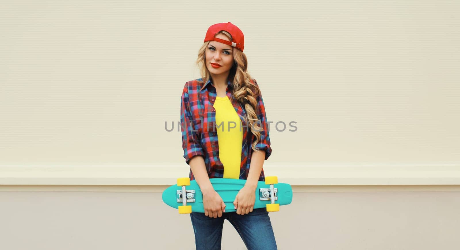 Portrait of stylish young blonde woman posing with skateboard on city street against white wall background