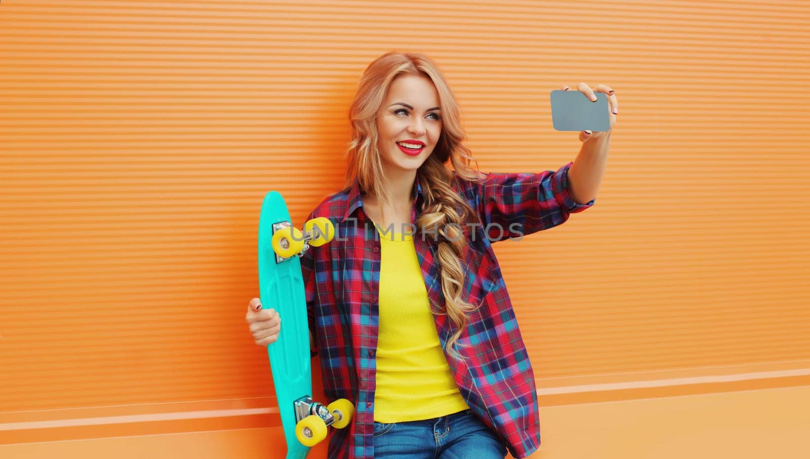 Summer portrait of happy smiling blonde young woman taking selfie with smartphone and skateboard on colorful orange background