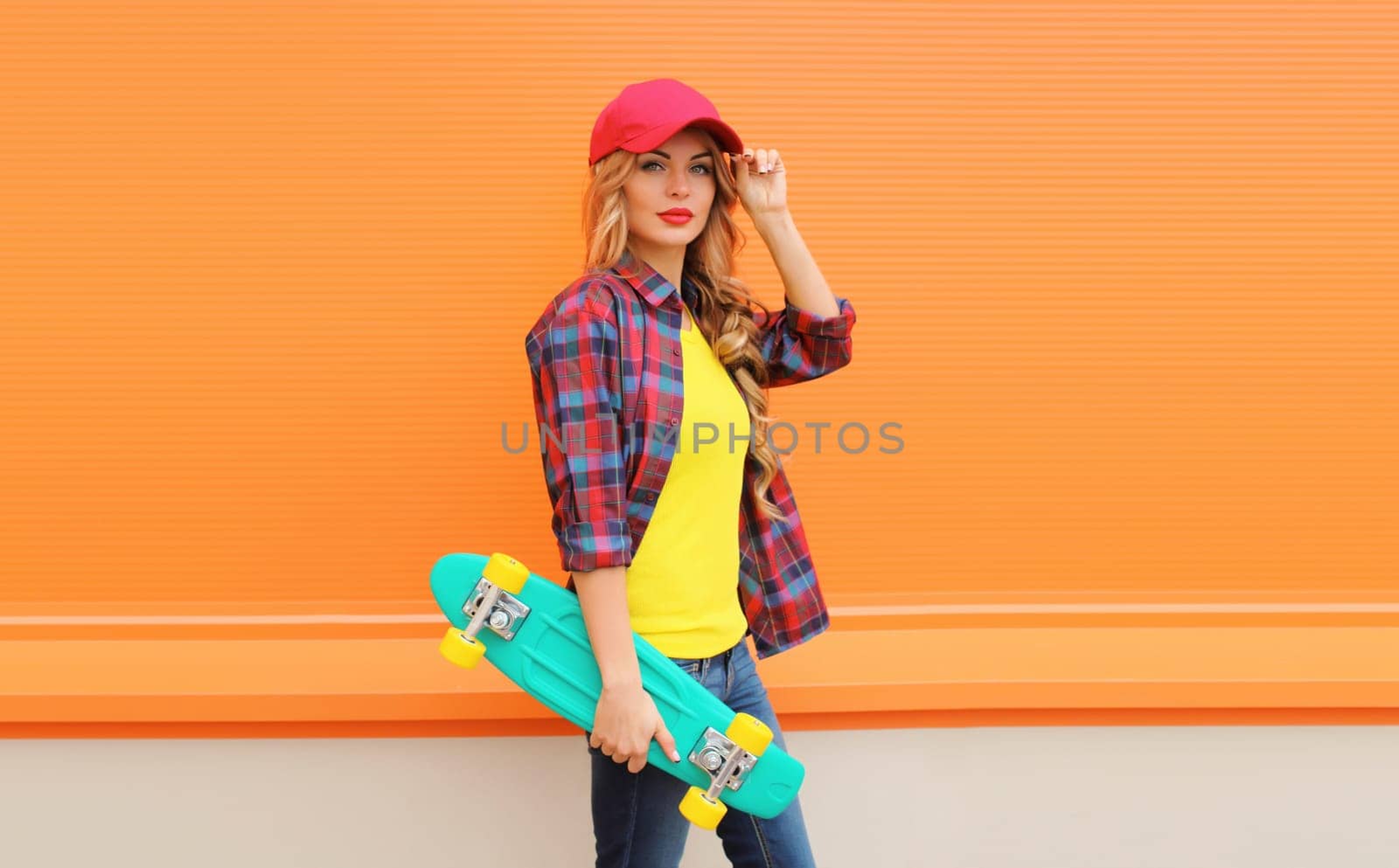 Portrait of stylish young blonde woman posing with skateboard in red baseball cap, shirt on city street against colorful orange background