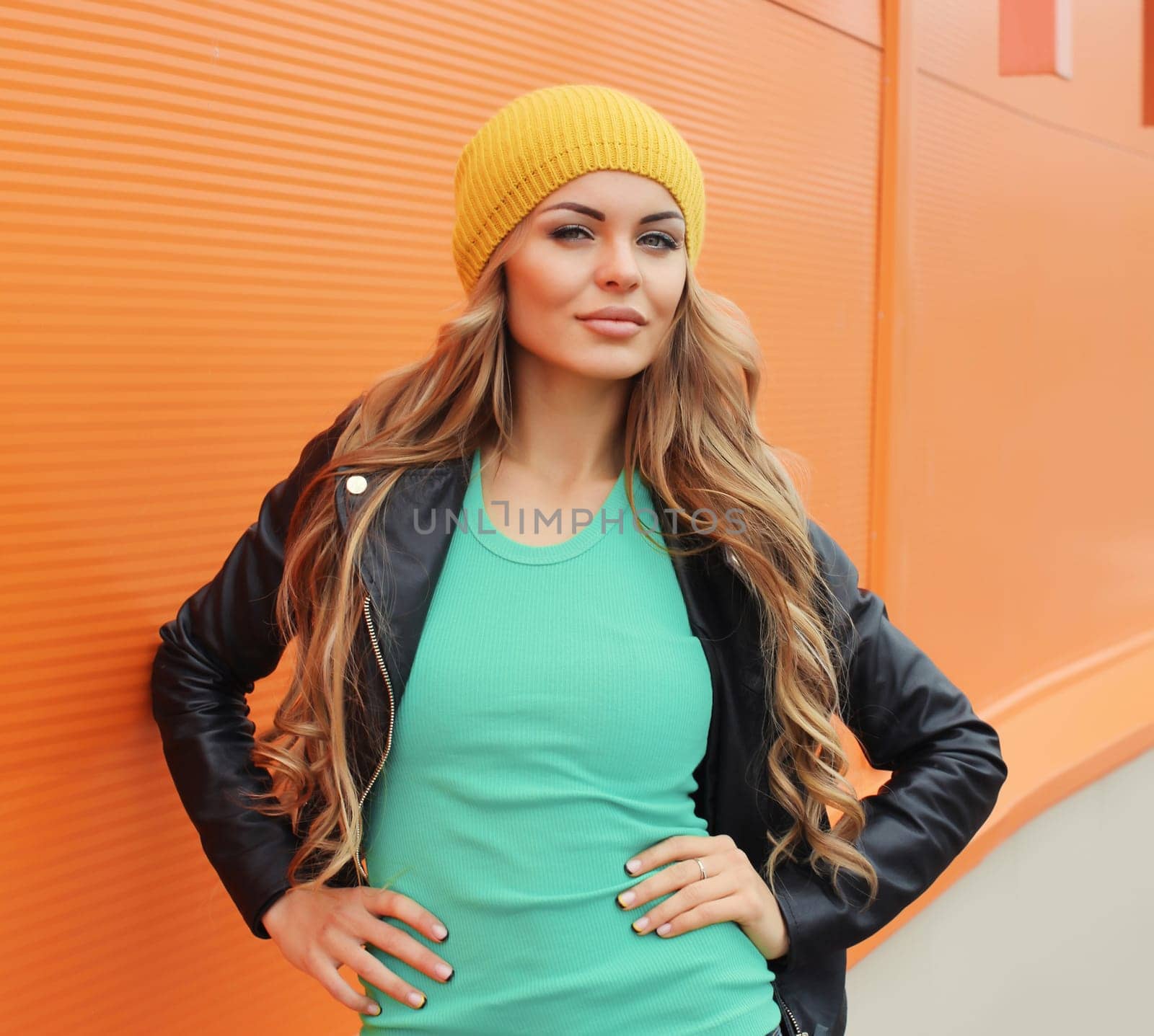 Portrait of beautiful young blonde woman posing in yellow hat, black leather jacket looking at camera on colorful orange background