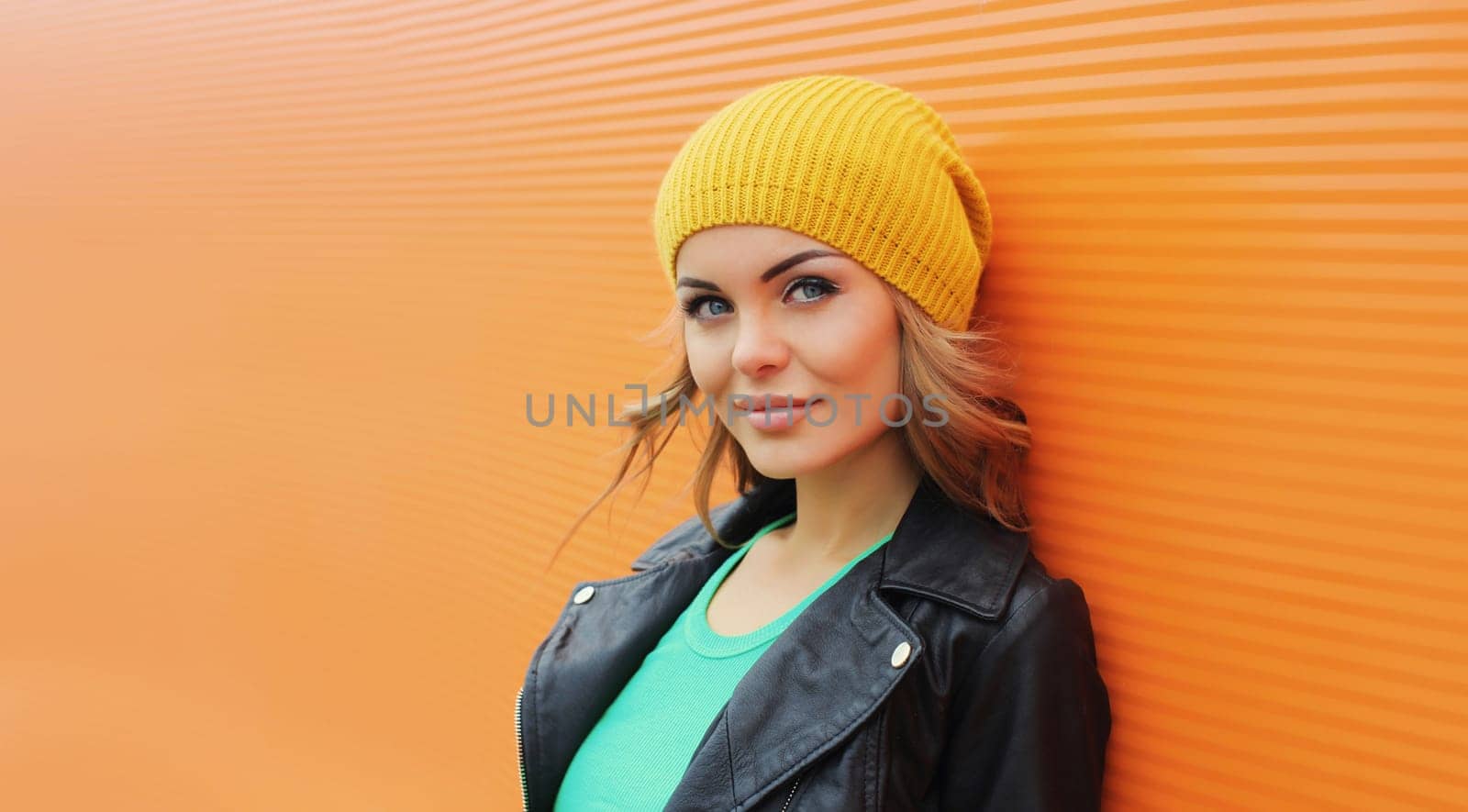 Portrait of beautiful young blonde woman posing in yellow hat, black leather jacket looking at camera on colorful orange background