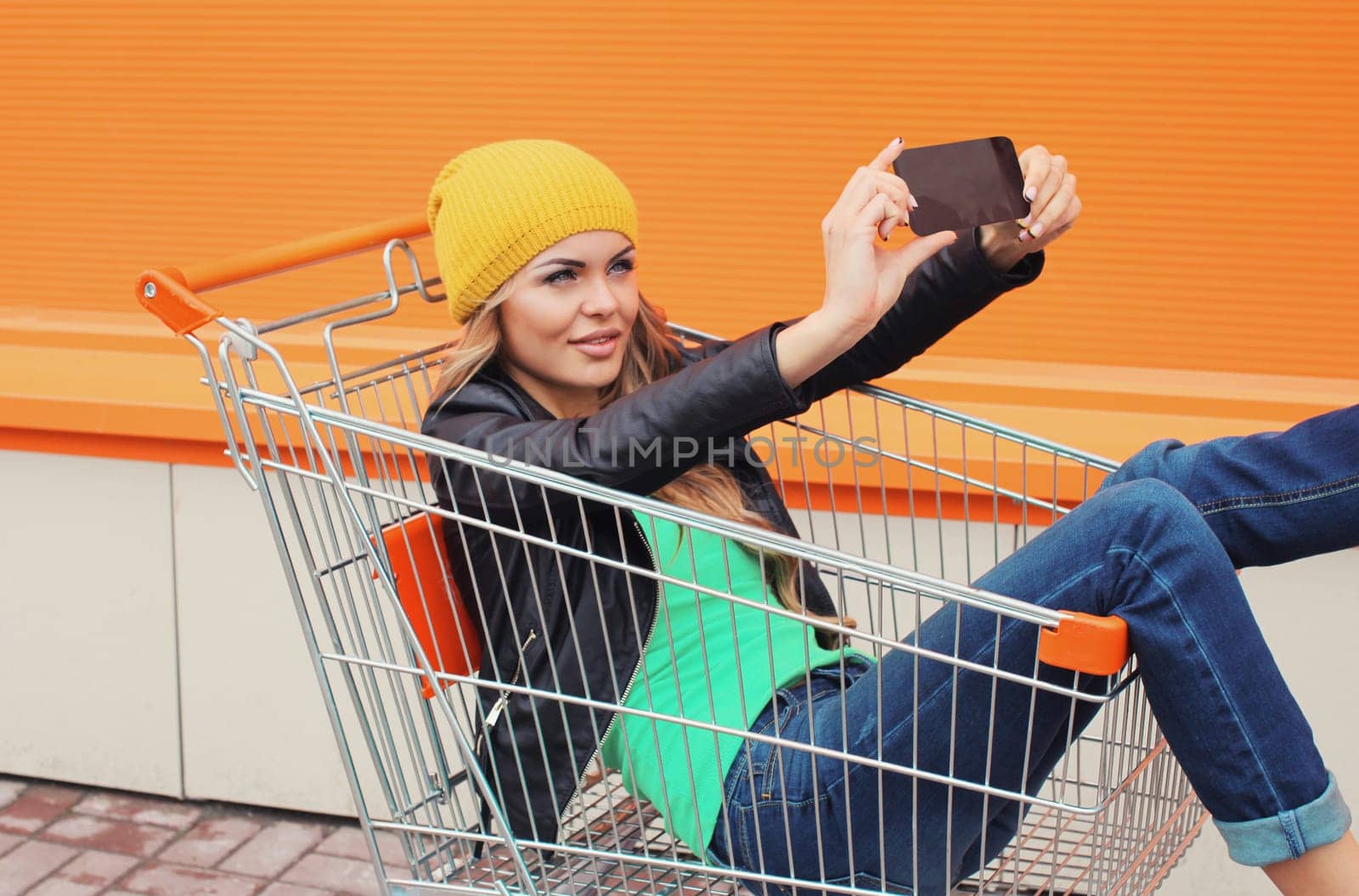 Stylish happy young blonde woman taking selfie with phone having fun sitting in shopping trolley cart in the city