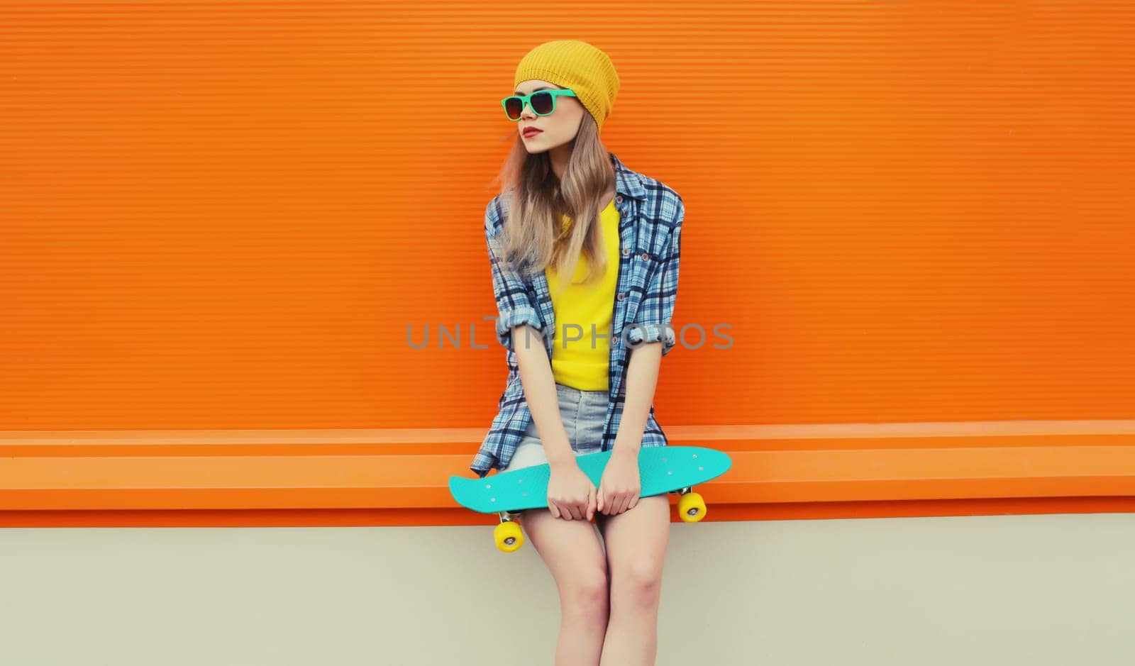 Summer portrait of stylish young woman with skateboard in colorful clothes, yellow hat looking away posing on orange background on city street