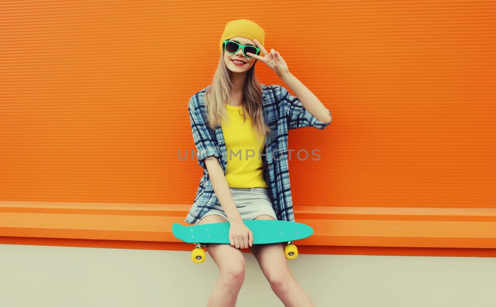 Summer portrait of happy cheerful stylish young woman with skateboard in colorful clothes, yellow hat posing on orange background on city street