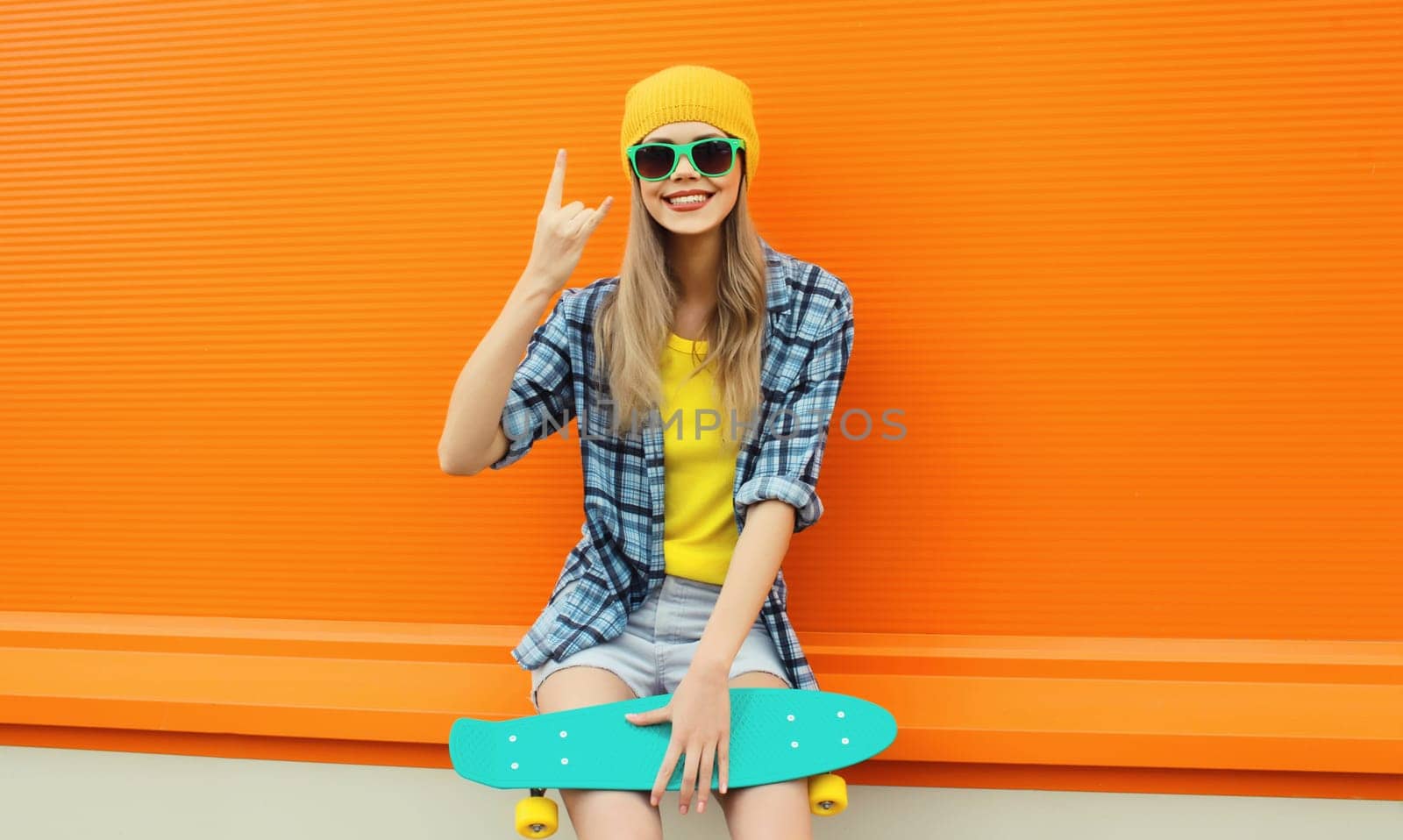 Summer portrait of happy cheerful stylish young woman with skateboard in colorful clothes, yellow hat posing on orange background on city street