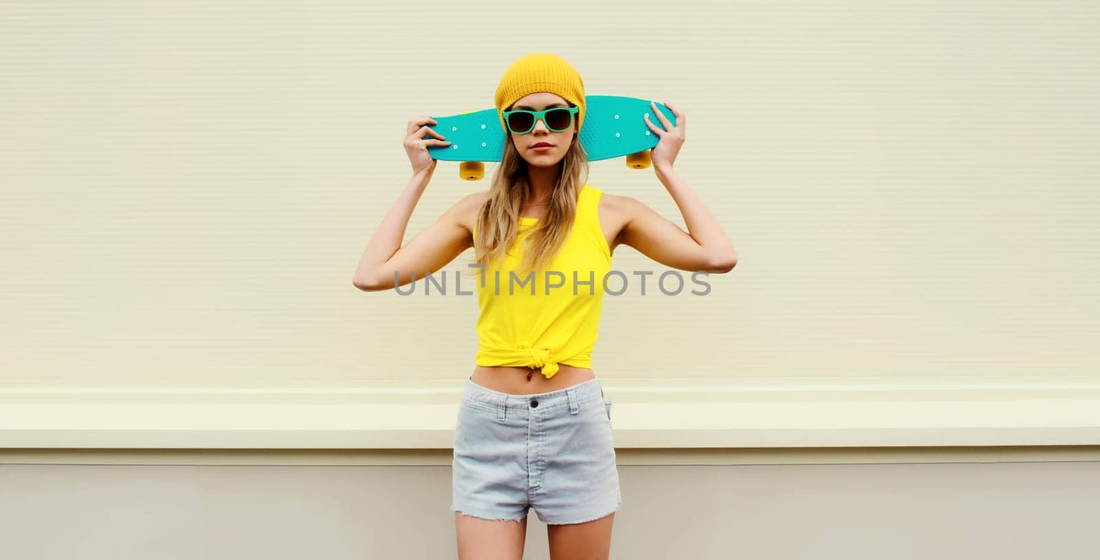Summer portrait of stylish blonde young woman with skateboard in colorful clothes, yellow hat posing on white background on city street