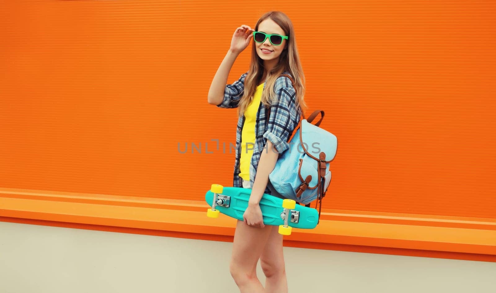 Summer portrait of happy cheerful stylish young woman with skateboard, backpack in colorful clothes posing on orange background on city street
