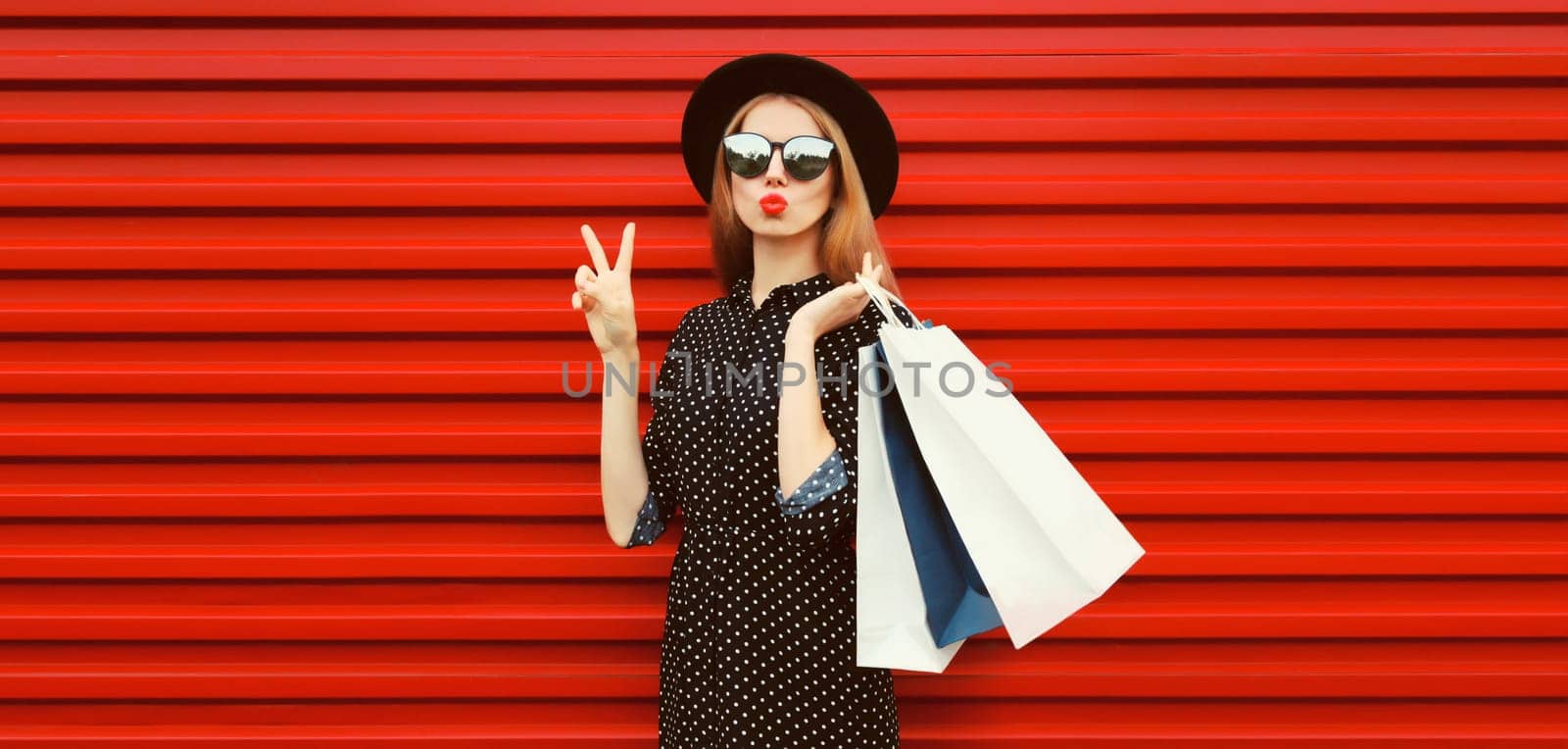 Portrait of beautiful young woman with shopping bags blowing her lips sends air kiss wearing black round hat on red background