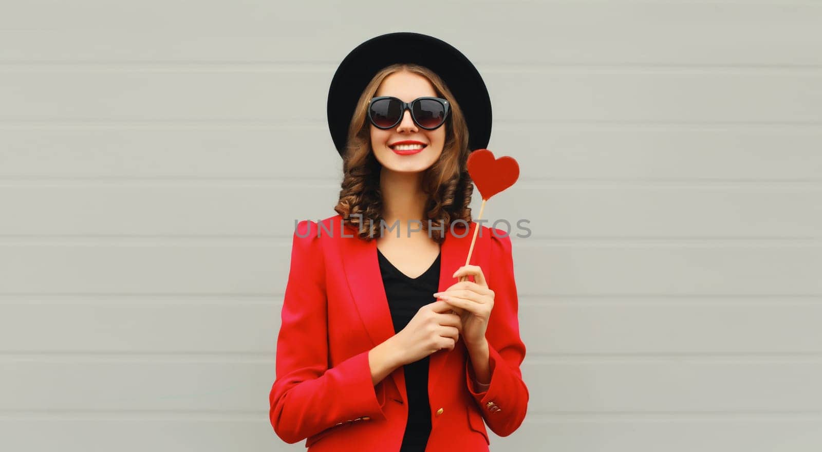 Beautiful happy woman with sweet red heart shaped lollipop on stick in black round hat