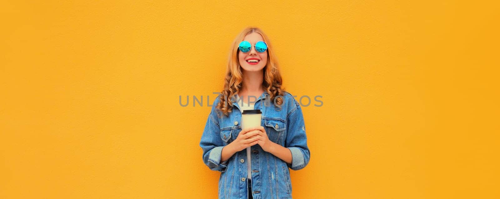 Beautiful happy smiling young woman with cup of coffee in denim jacket on yellow studio background