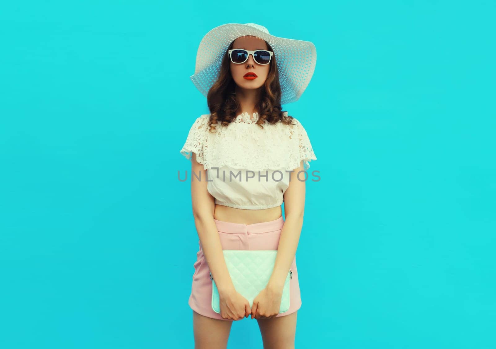 Beautiful caucasian young woman model posing with handbag clutch wearing white summer straw hat on blue studio background