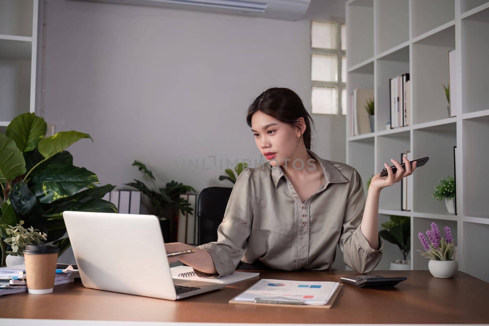 Young Asian business woman sits on the phone in an online business meeting using a laptop in a modern home office decorated with shady green plants..