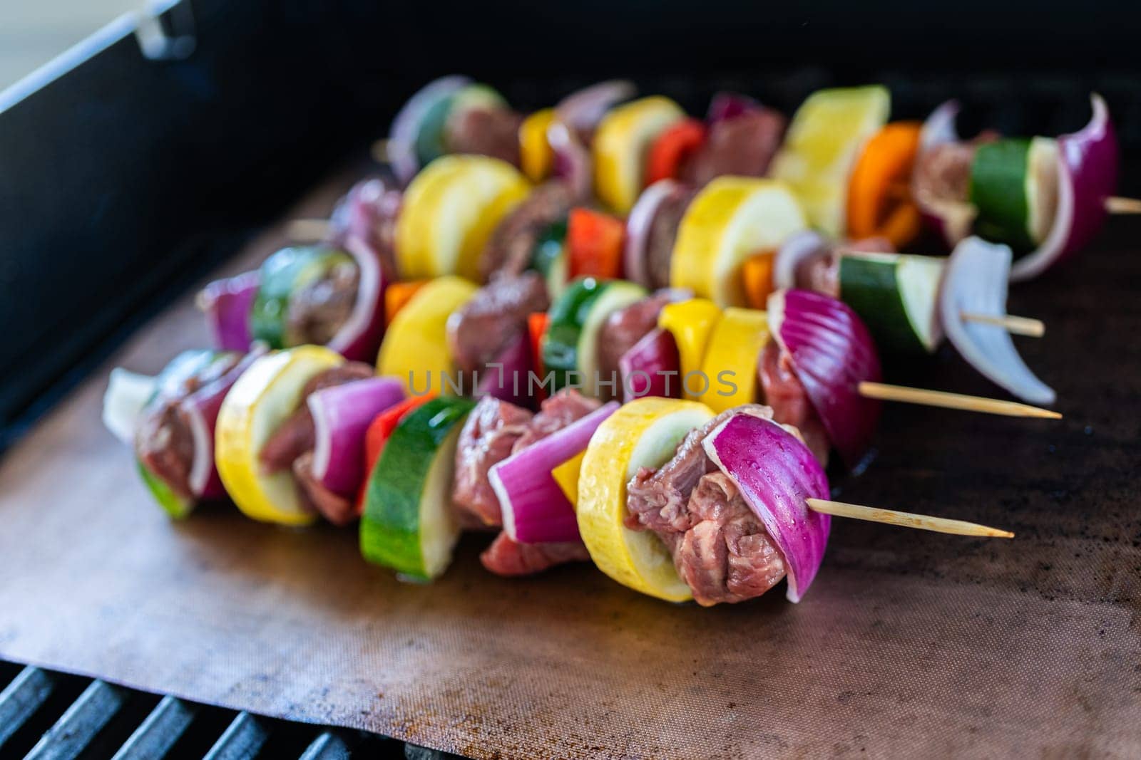 Skewered beef and fresh veggies sizzle on a copper grill mat over a gas outdoor grill.
