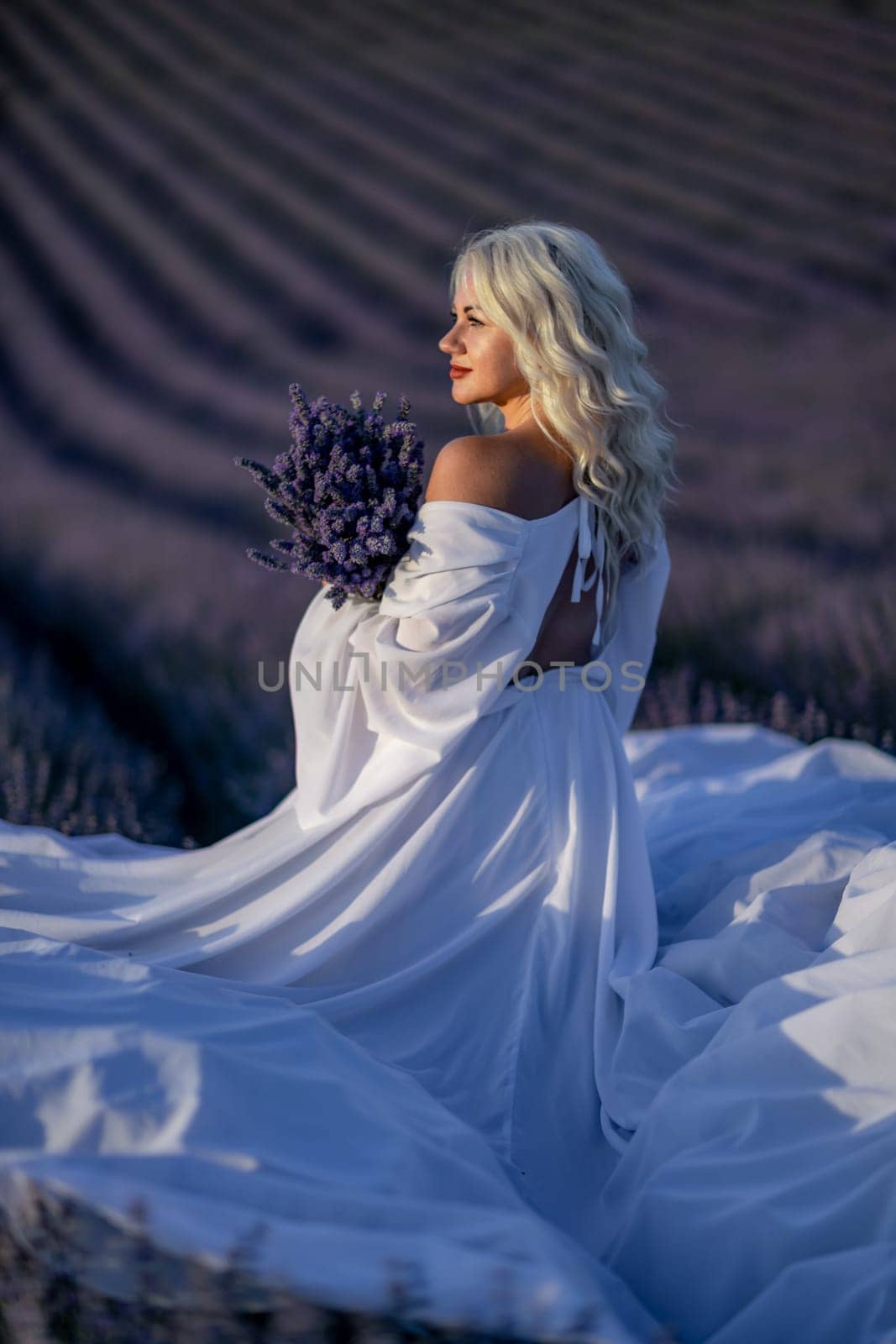 Blonde woman poses in lavender field at sunset. Happy woman in white dress holds lavender bouquet. Aromatherapy concept, lavender oil, photo session in lavender by Matiunina