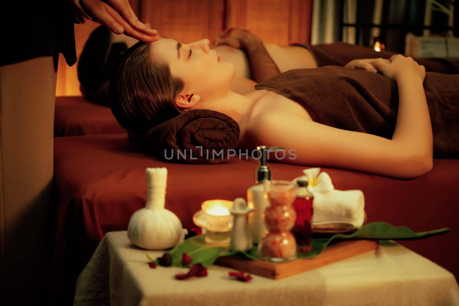 Couple customer enjoying relaxing anti-stress head massage and pampering facial beauty skin recreation leisure in warm candle lighting ambient salon spa in luxury resort or hotel. Quiescent