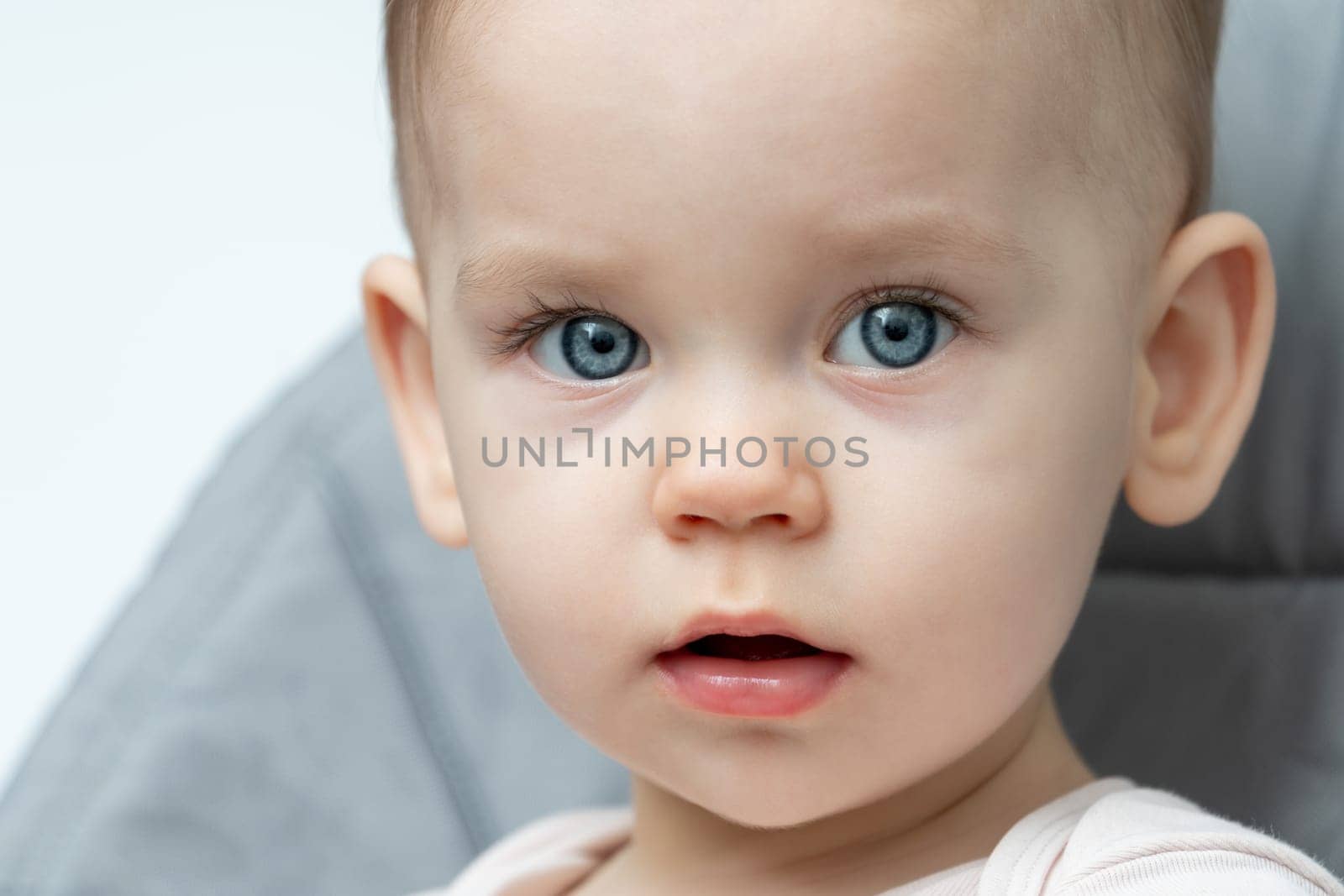 A happy baby with blue eyes is sitting in a high chair, smiling at the camera while showing off their cute nose, cheek, eyelash, ear, and iris