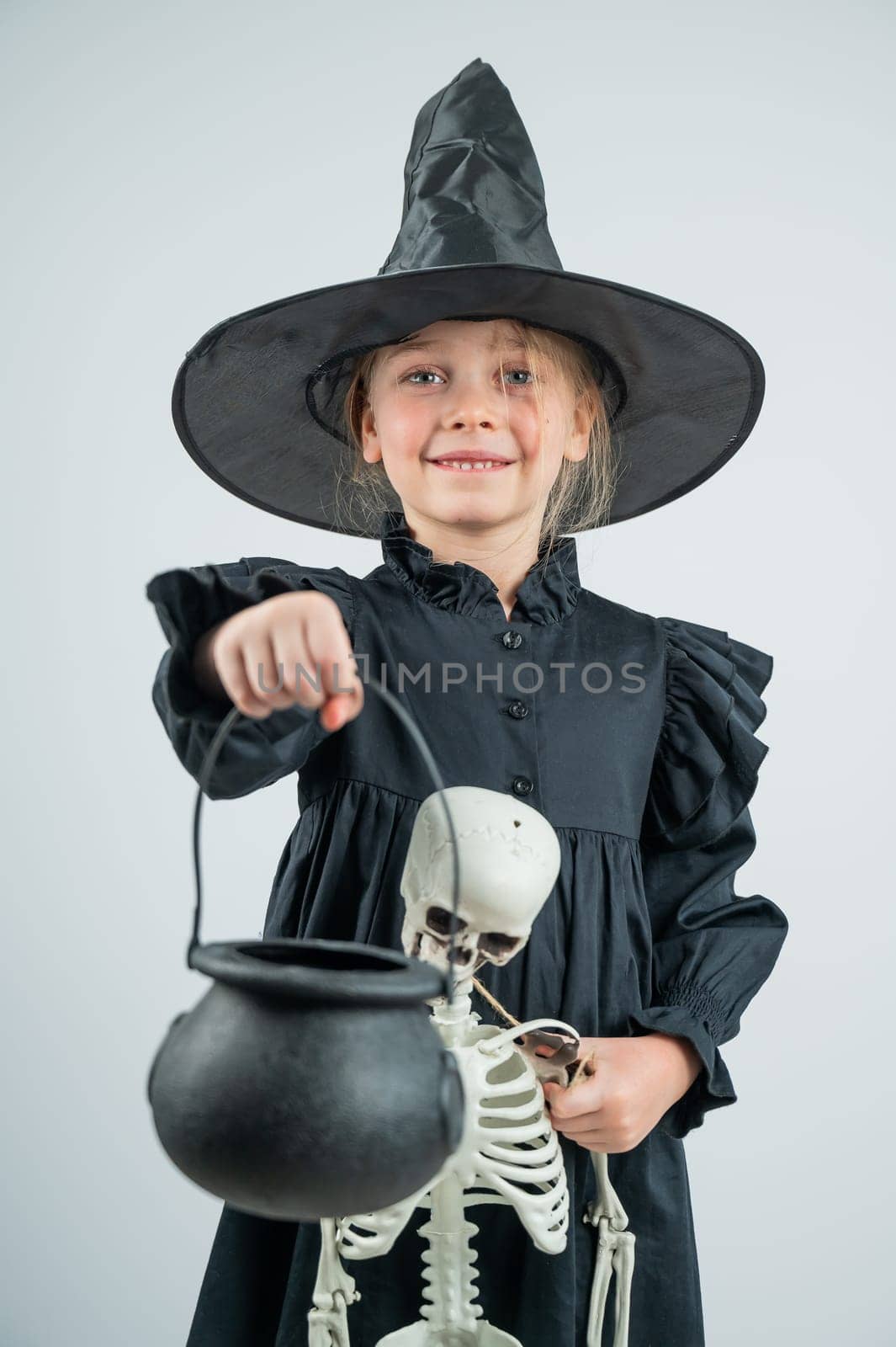 Portrait of a little Caucasian girl in a witch costume holding a cauldron and a skeleton on a white background. Vertical photo. by mrwed54
