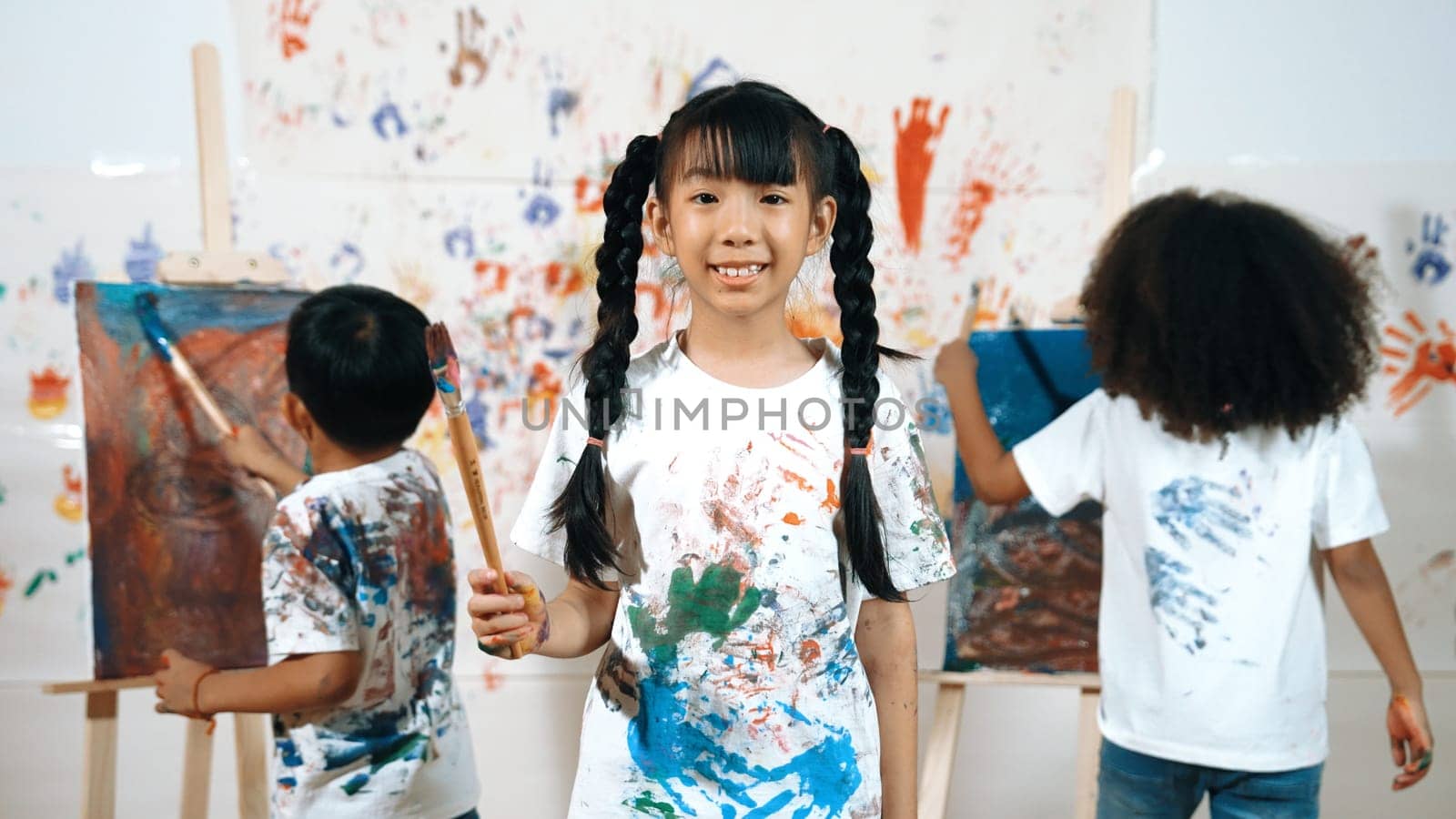 Creative girl pose at camera while diverse children painting behind. Erudition. by biancoblue