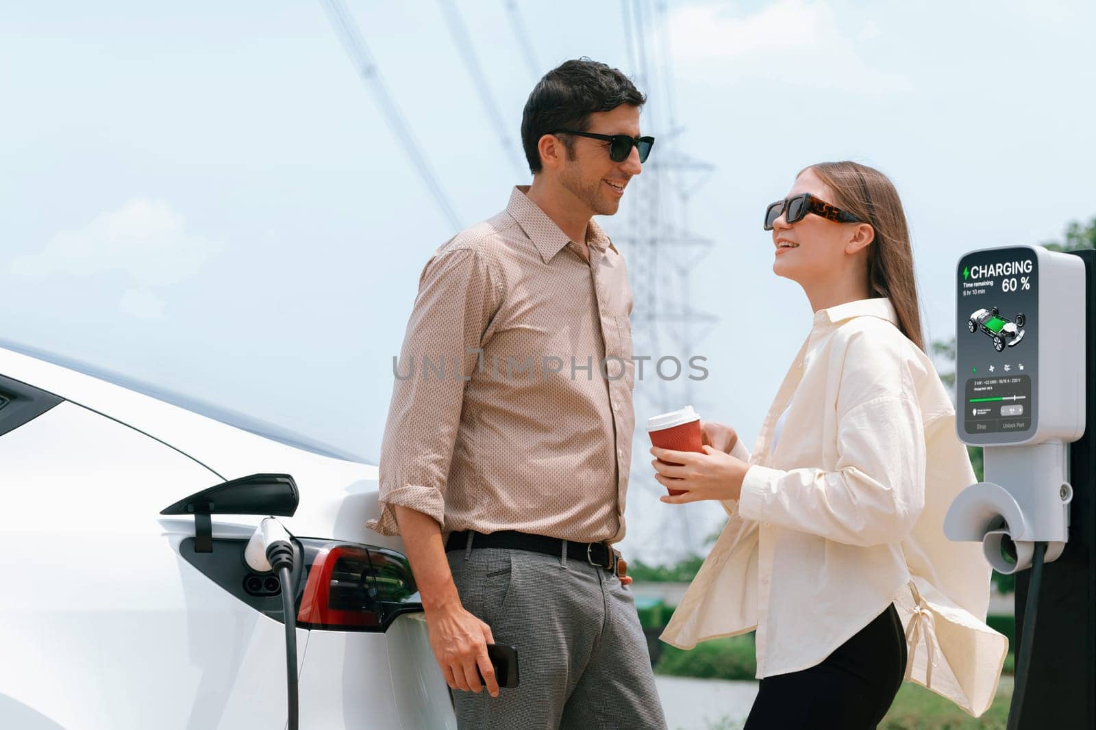 Young couple with coffee recharge EV car battery at charging station connected to electrical industrial power grid tower. Couple with shopping and travel using eco electric car lifestyle. Expedient