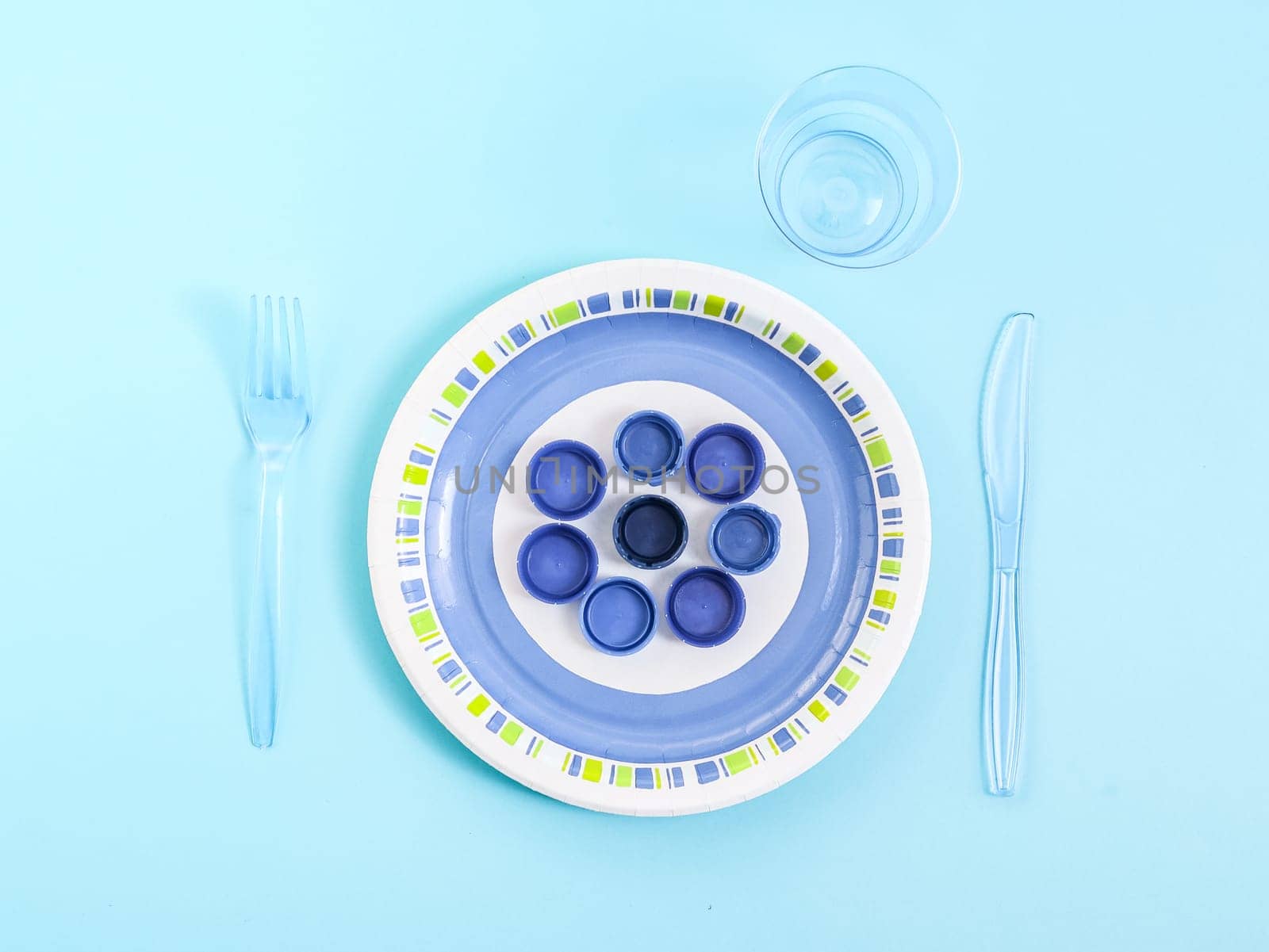 A paper plate with plastic bottle caps, with a plastic glass, a fork and a knife on a light blue background, a flat lay close-up. The concept of ecology, disposable cutlery, serving and plastic food.